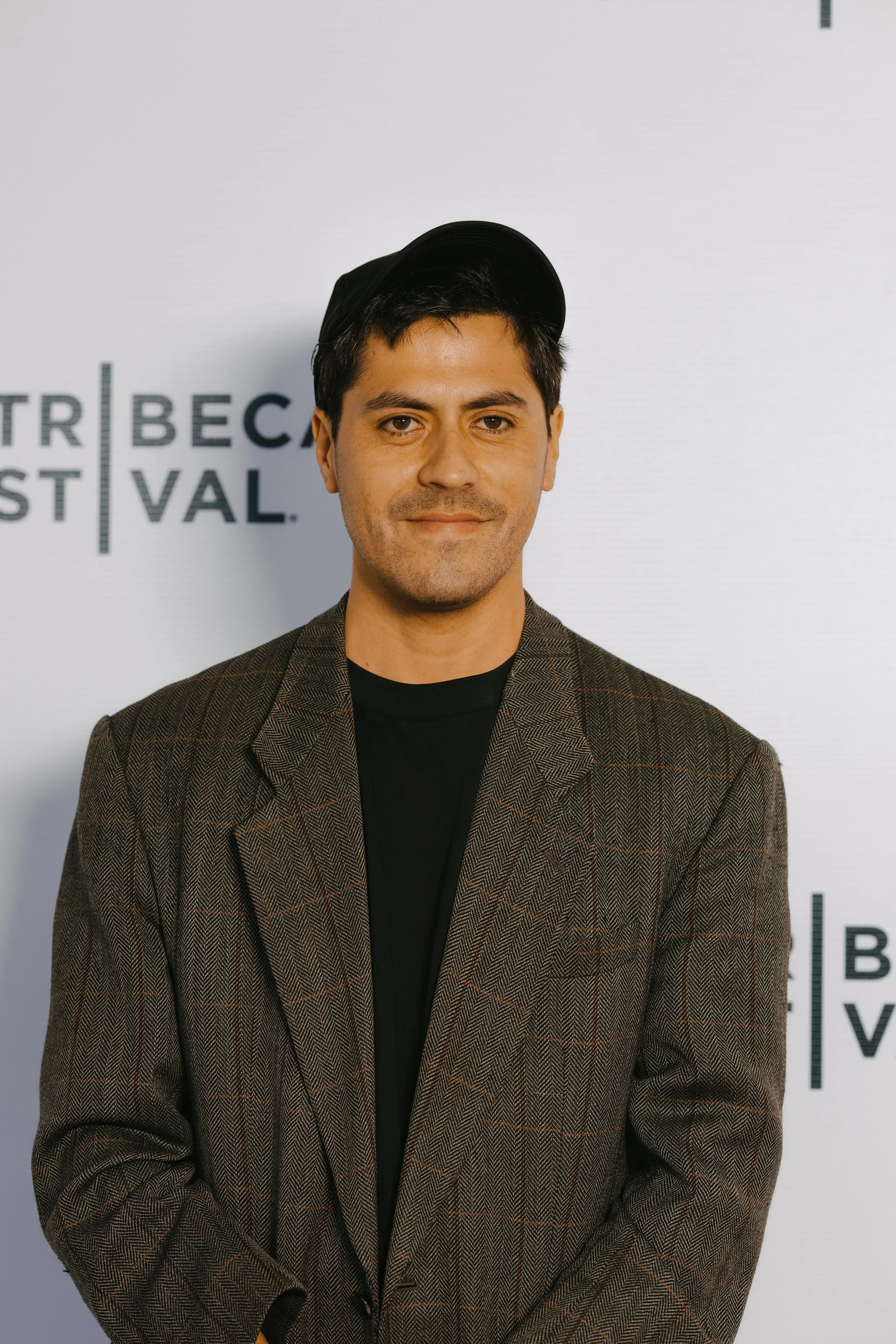 A young man with dark hair, wearing a black cap, a black shirt, and a brown plaid blazer, stands in front of a backdrop with partially visible text, smiling subtly.