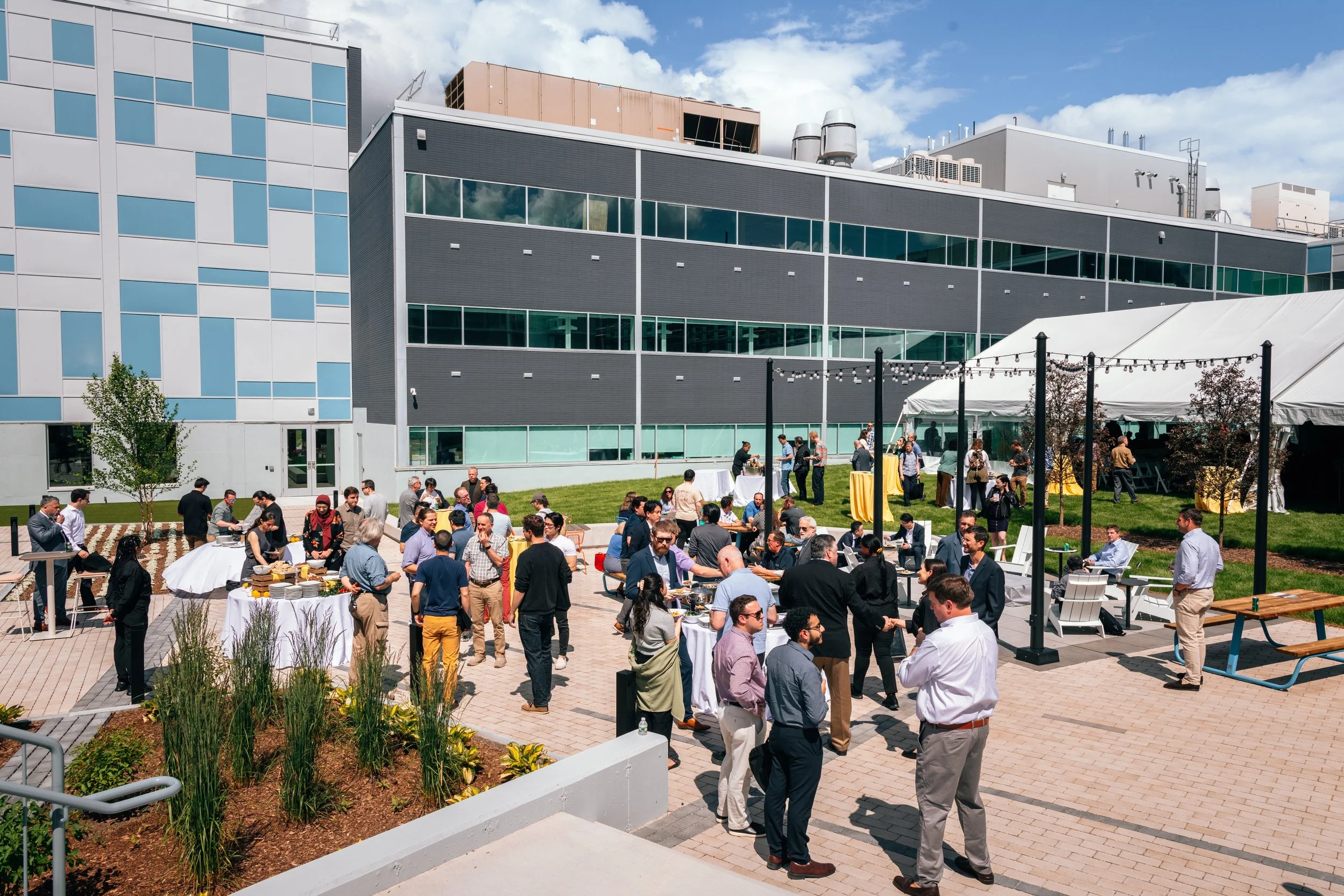 Outdoor corporate event or wedding reception with people mingling, eating, and talking under a white tent on a paved patio area in front of a modern office building under a partly cloudy sky.