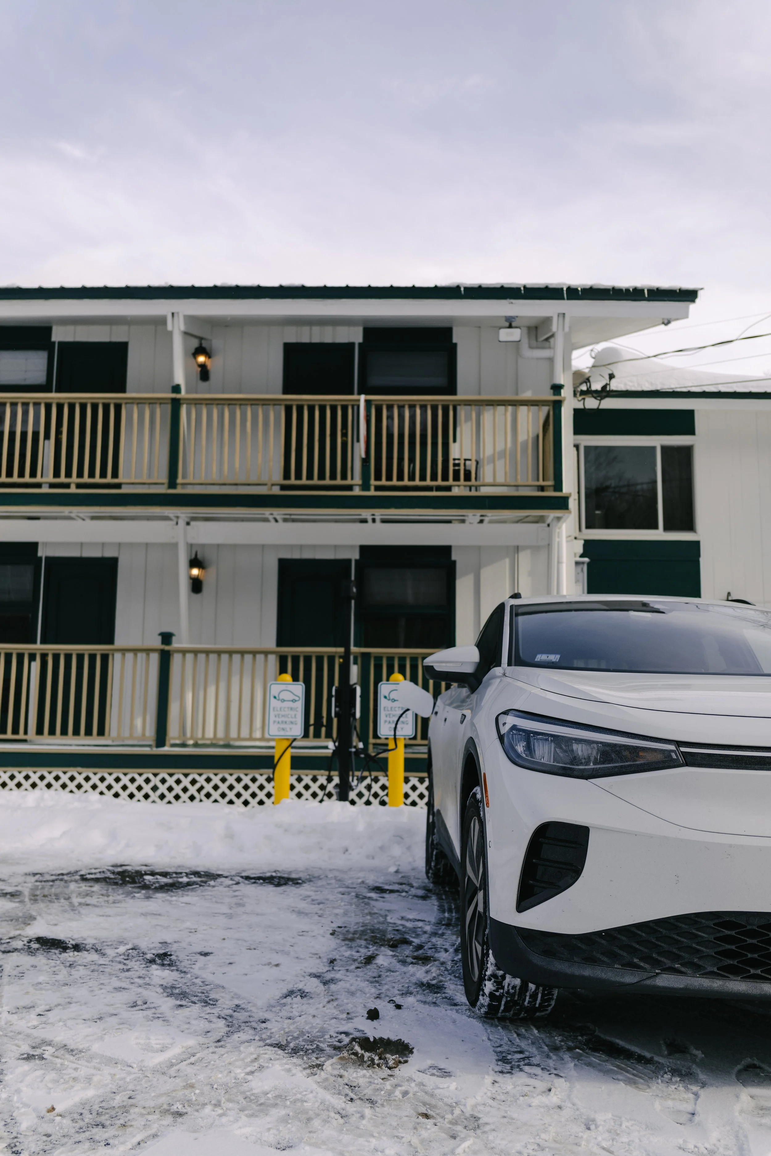 A white electric vehicle is parked at a charging station in front of a multi-story building with balconies and a snowy ground.