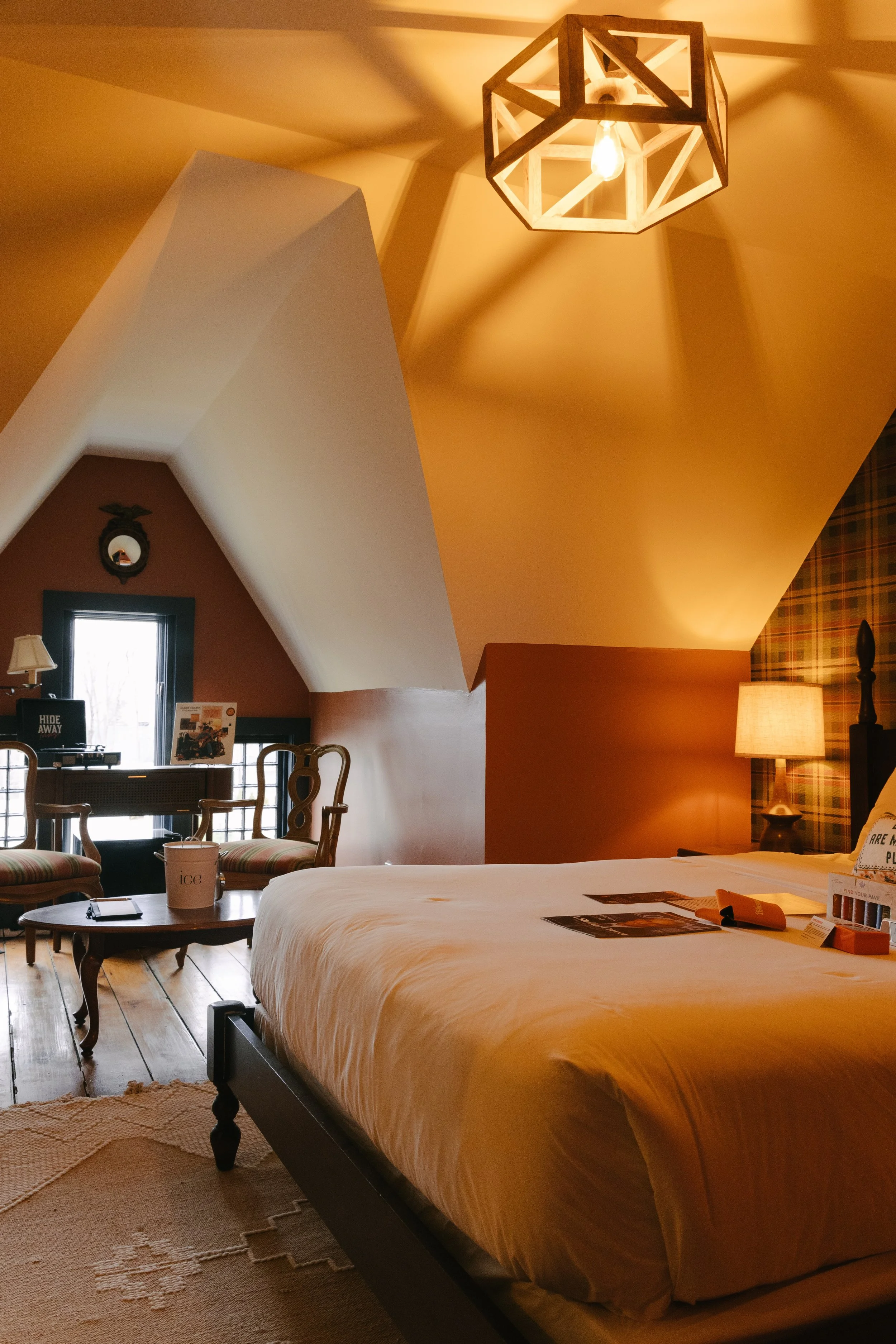 A cozy hotel room with a large bed, wooden furniture, a lamp, and a window with sunlight. The ceiling has angled walls and a geometric light fixture.