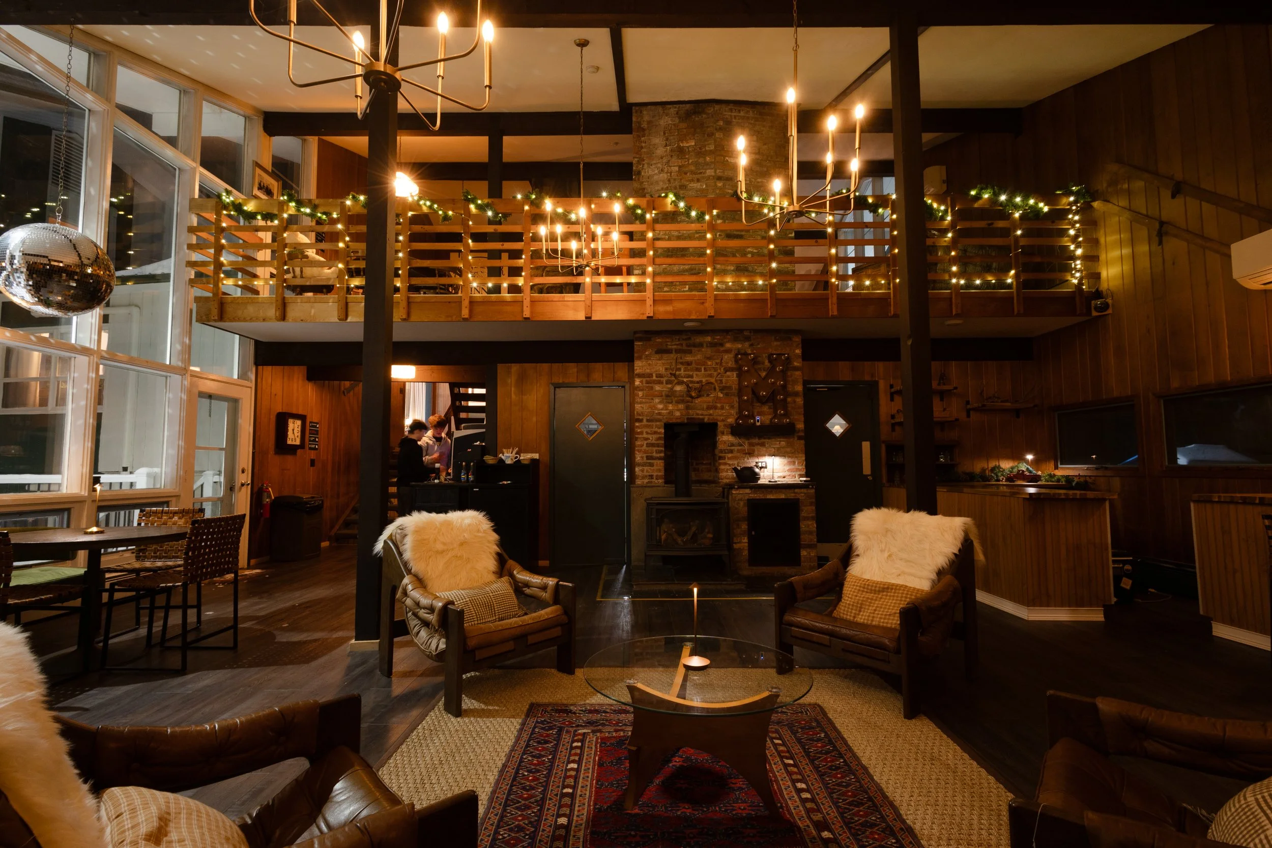 Indoor lounge area with four leather armchairs with pillows and faux fur throws around a glass coffee table, a rug, and a lit candle. In the background, a brick chimney with a fireplace, a bar with a couple of people, and a two-story space with a woo