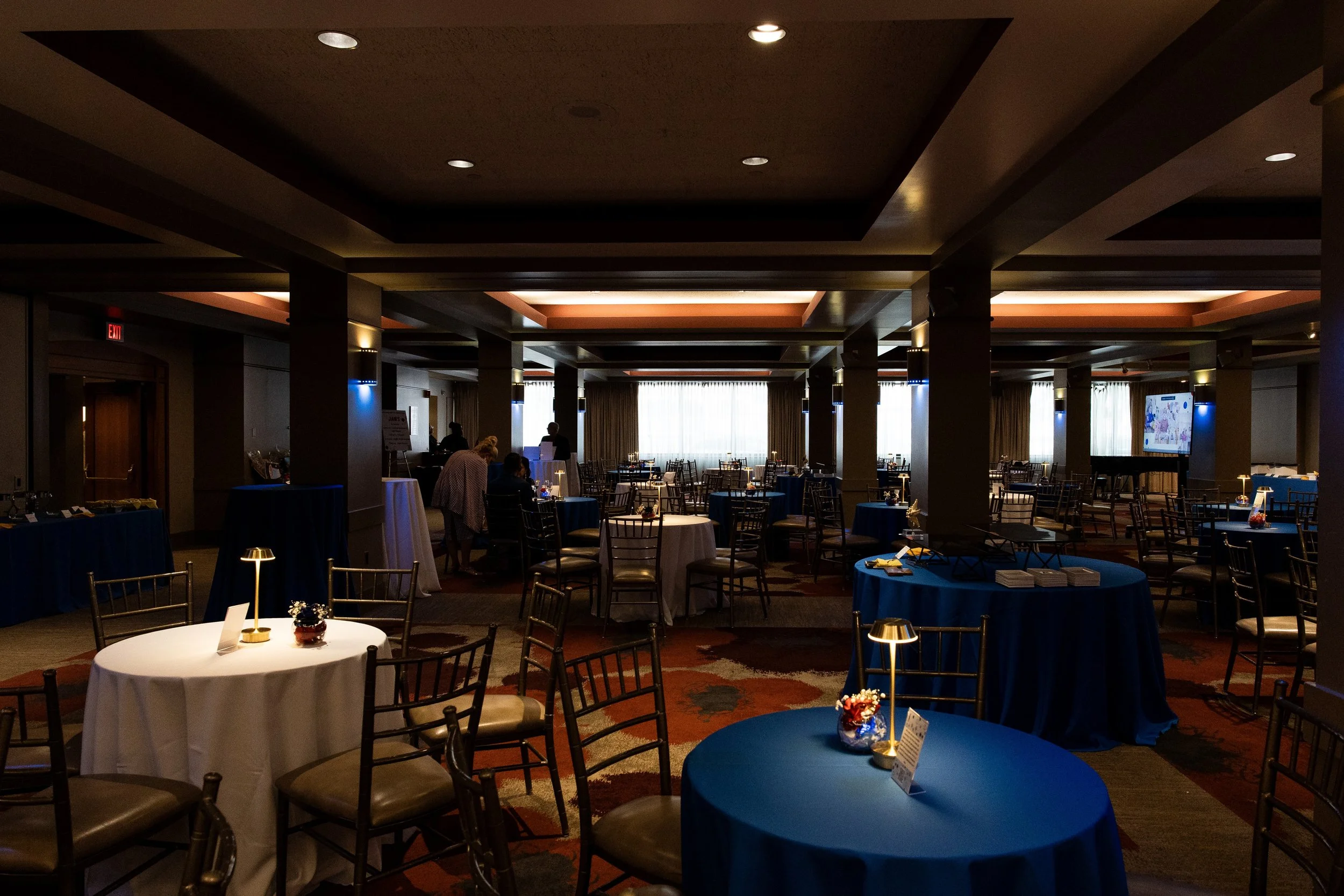 A banquet hall with round tables covered in white and blue tablecloths, decorated with small floral centerpieces and table numbers. The room has a high ceiling with recessed lighting and large windows with curtains, and a few people are arranging or 