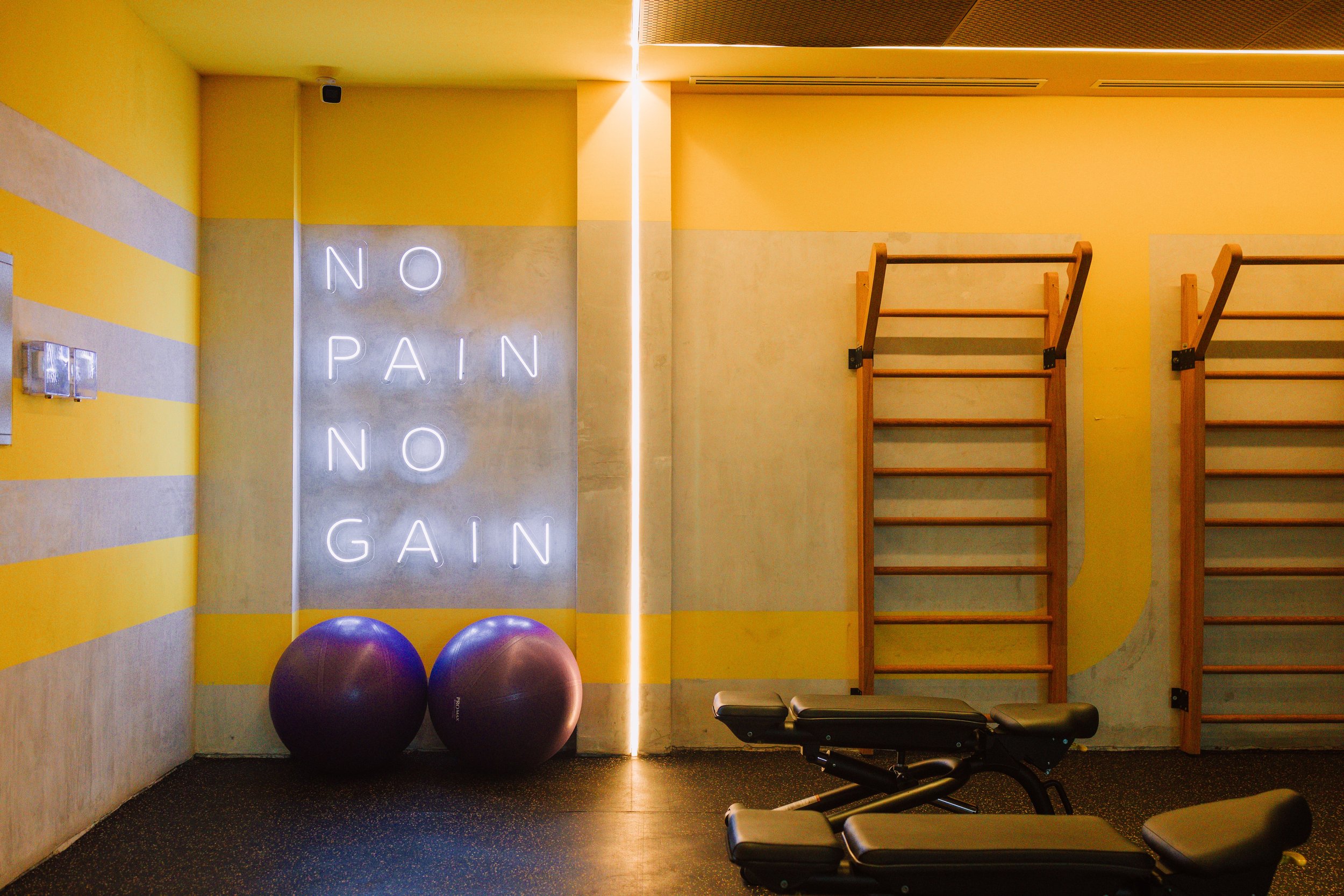 Interior of a gym or fitness studio with yellow striped walls, neon sign that says 'NO PAIN NO GAIN', exercise balls, and wooden wall bars