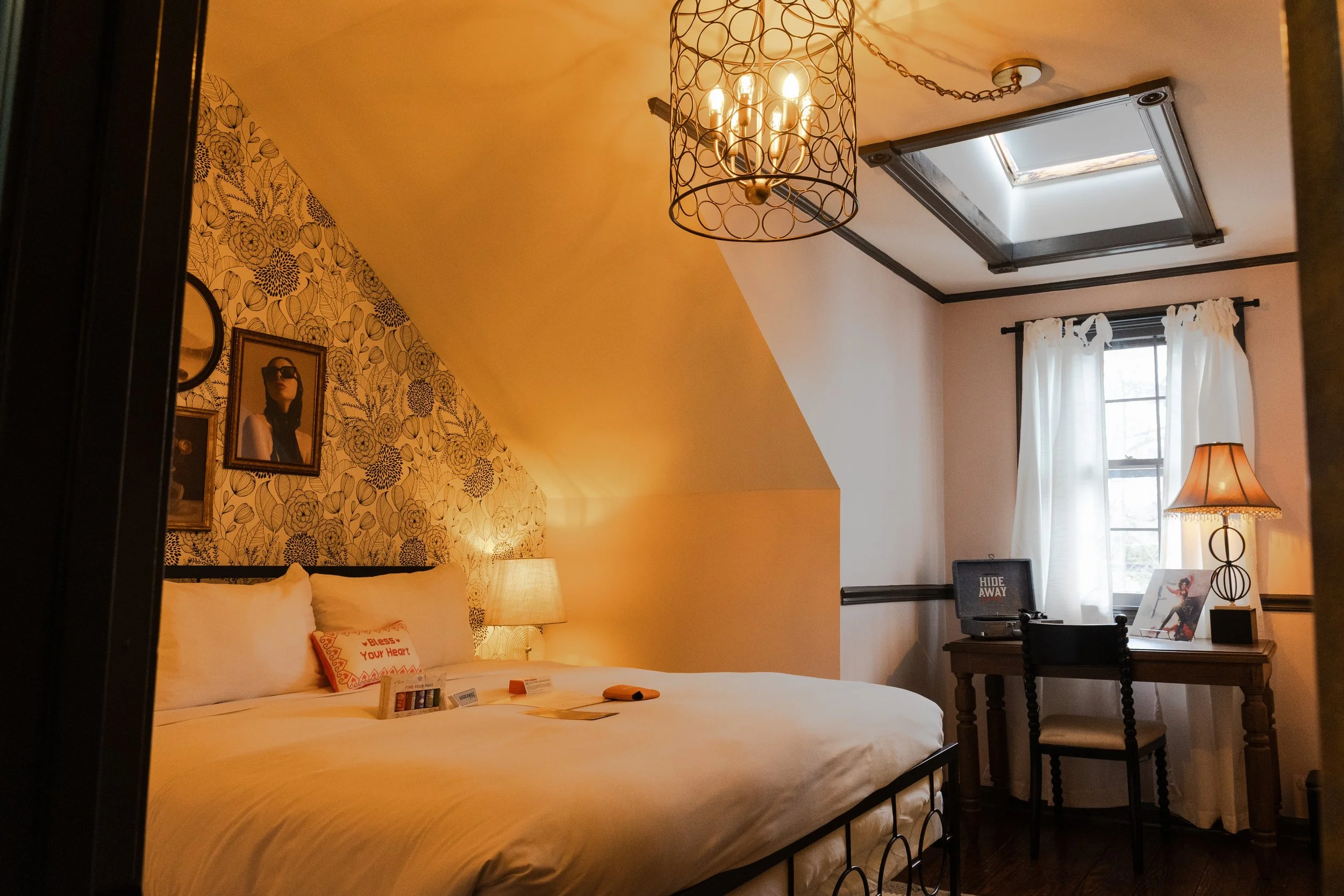 Cozy bedroom with a large bed, patterned floral wallpaper, framed photos, a bedside lamp, a desk with a computer, a table lamp, a window with white curtains, and a skylight.