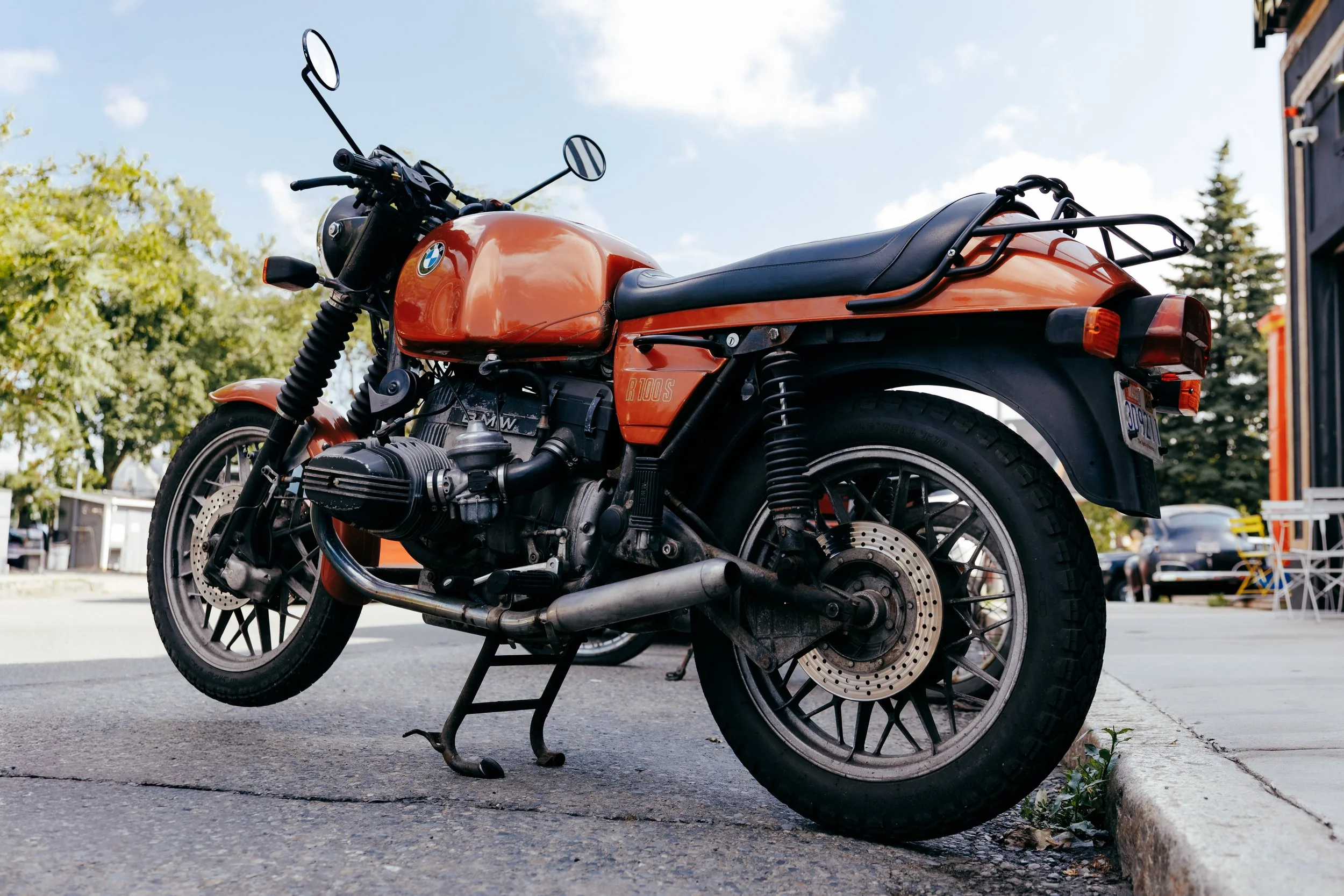 An orange BMW motorcycle parked on a city street curb, viewed from the side under a partly cloudy sky, with trees and buildings in the background.