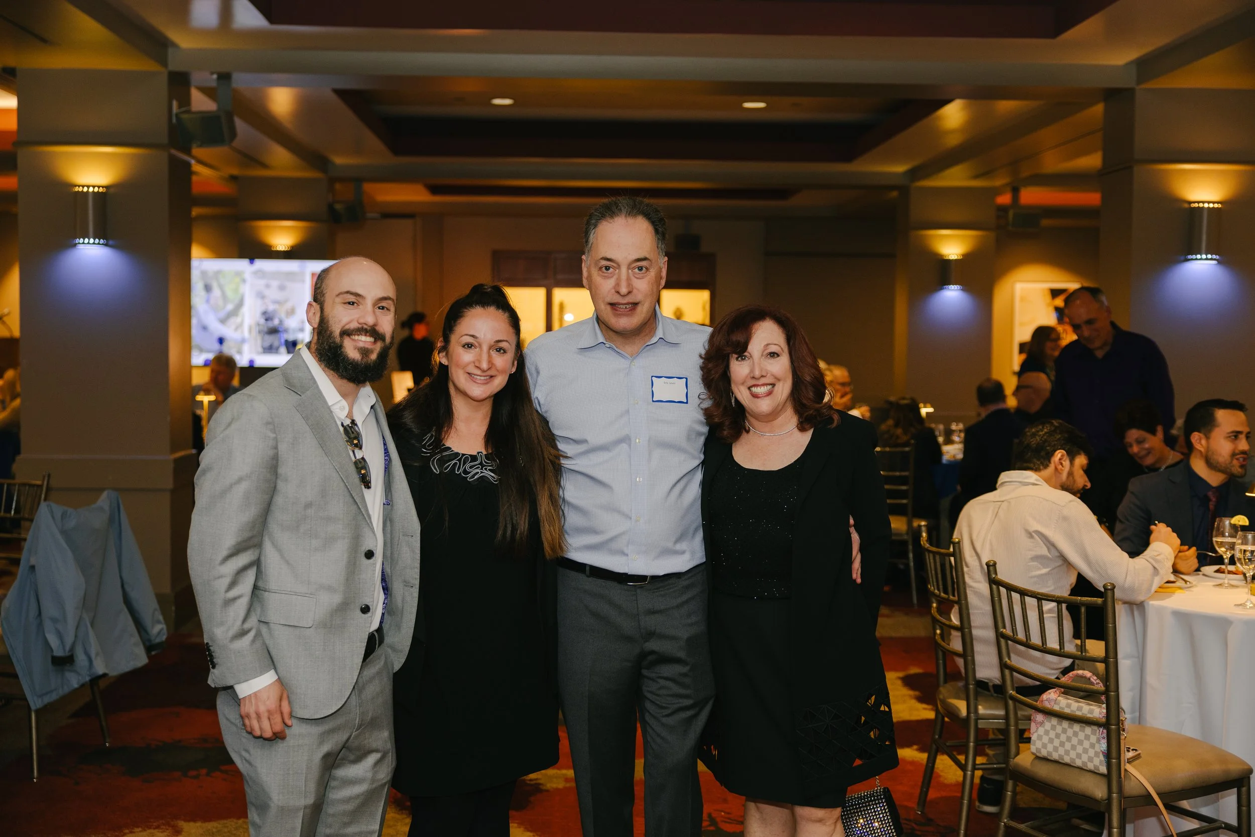 Four people standing together at a formal gathering, smiling, with a crowd of seated and standing people in the background. The setting appears to be a banquet or conference room with warm lighting and decorated tables.