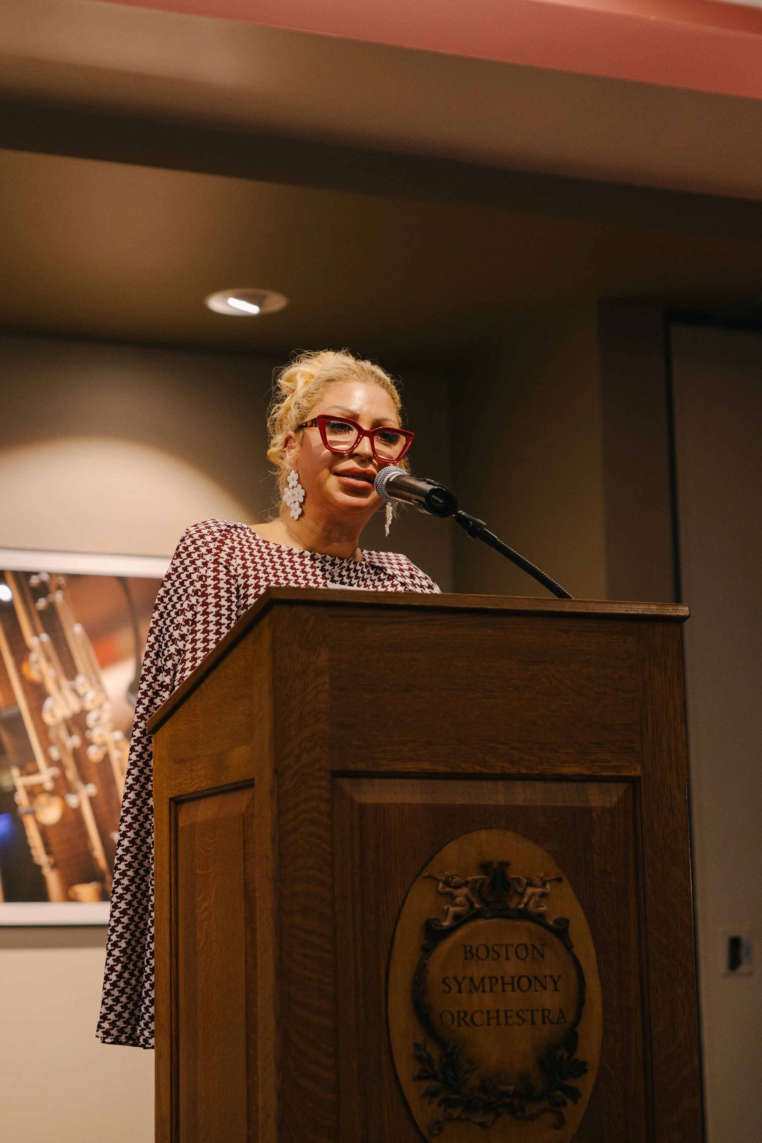 A woman with blonde hair, red glasses, and floral earrings speaking at a podium with the Boston Symphony Orchestra emblem, in a room with warm lighting and musical instruments in the background.