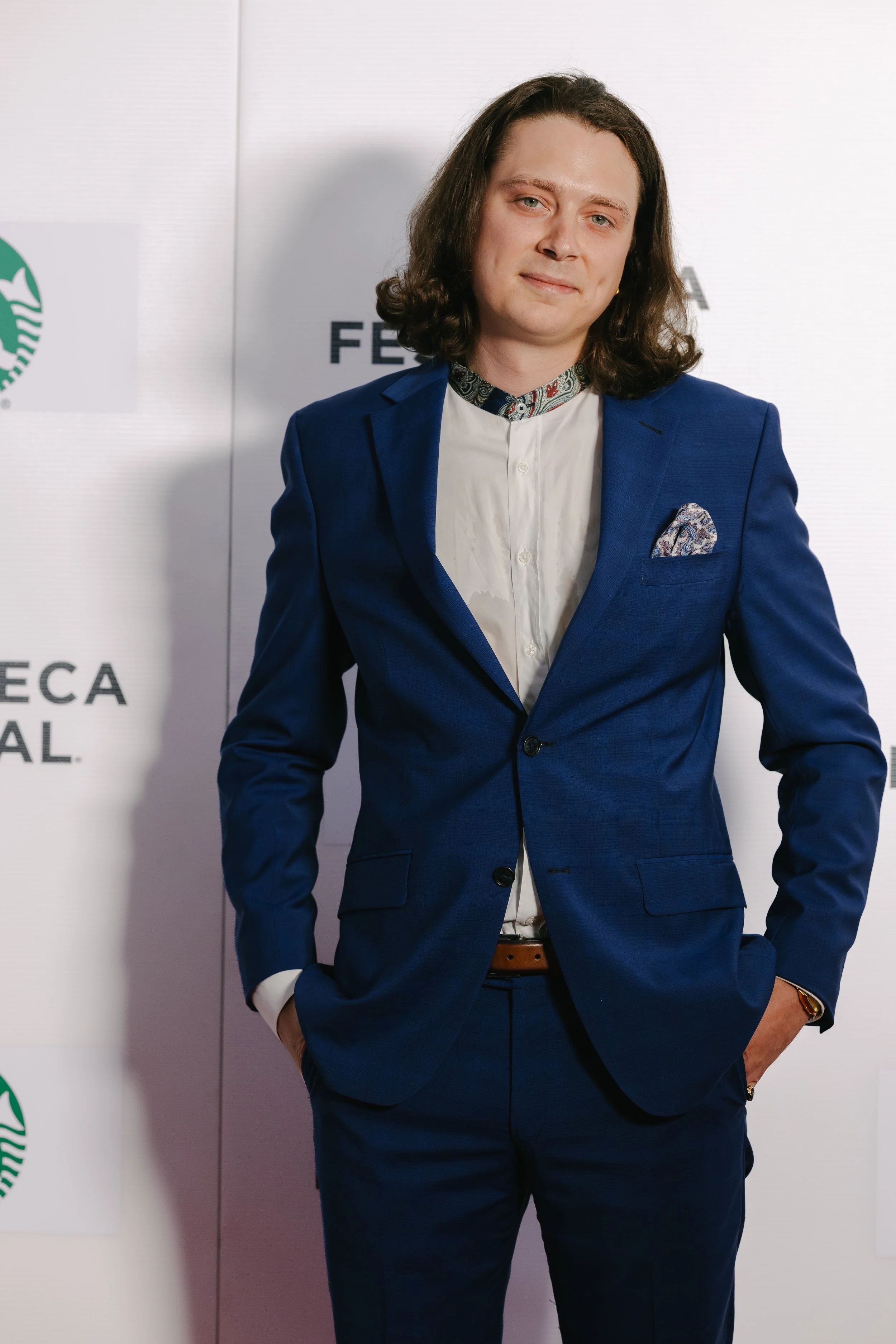 A man with shoulder-length dark hair, wearing a blue suit with a white shirt, standing with hands in pockets at an event, with visible logos and text in the background.