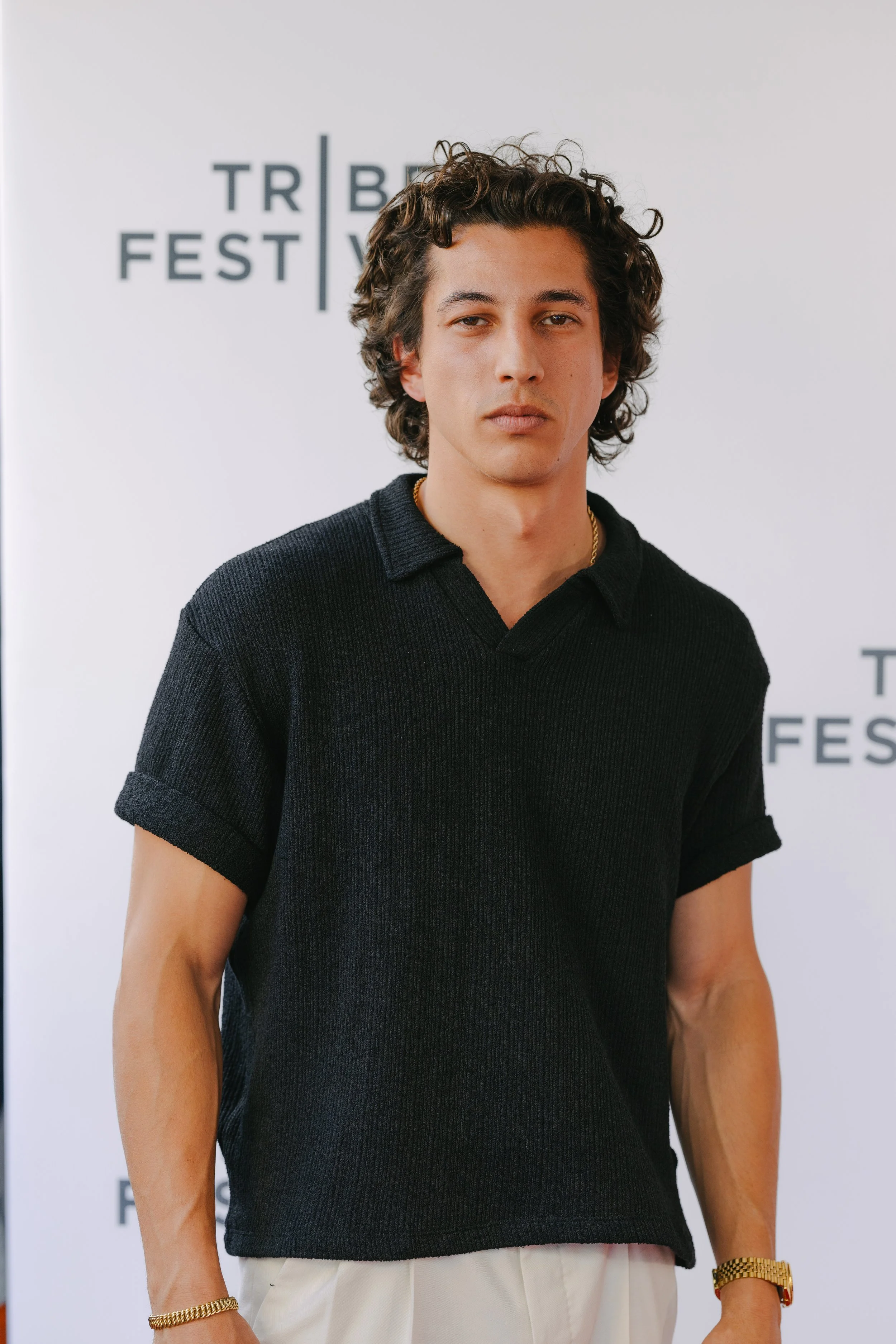 A young man with curly hair wearing a black short-sleeve polo shirt and beige pants at a film festival.