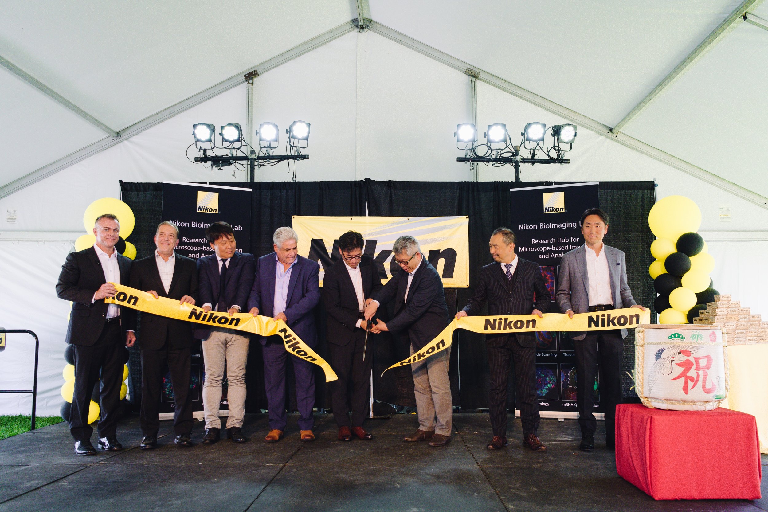 Group of people at a Nikon company event participating in a ribbon-cutting ceremony under a large tent, with Nikon banners and yellow and black balloon decorations.