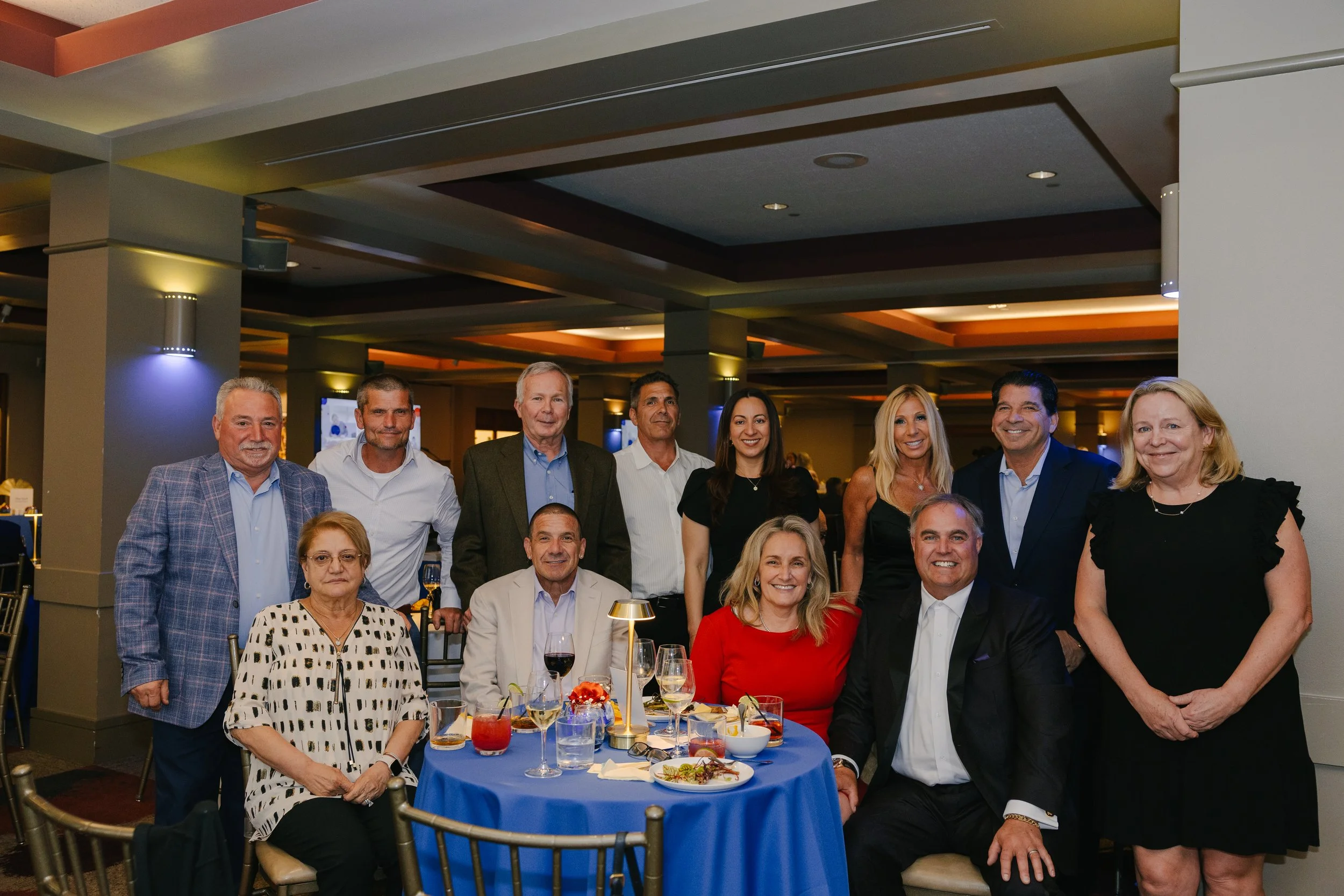 Group of ten people, some standing and some sitting, gathered at a table during a celebration or formal event in a decorated banquet hall.