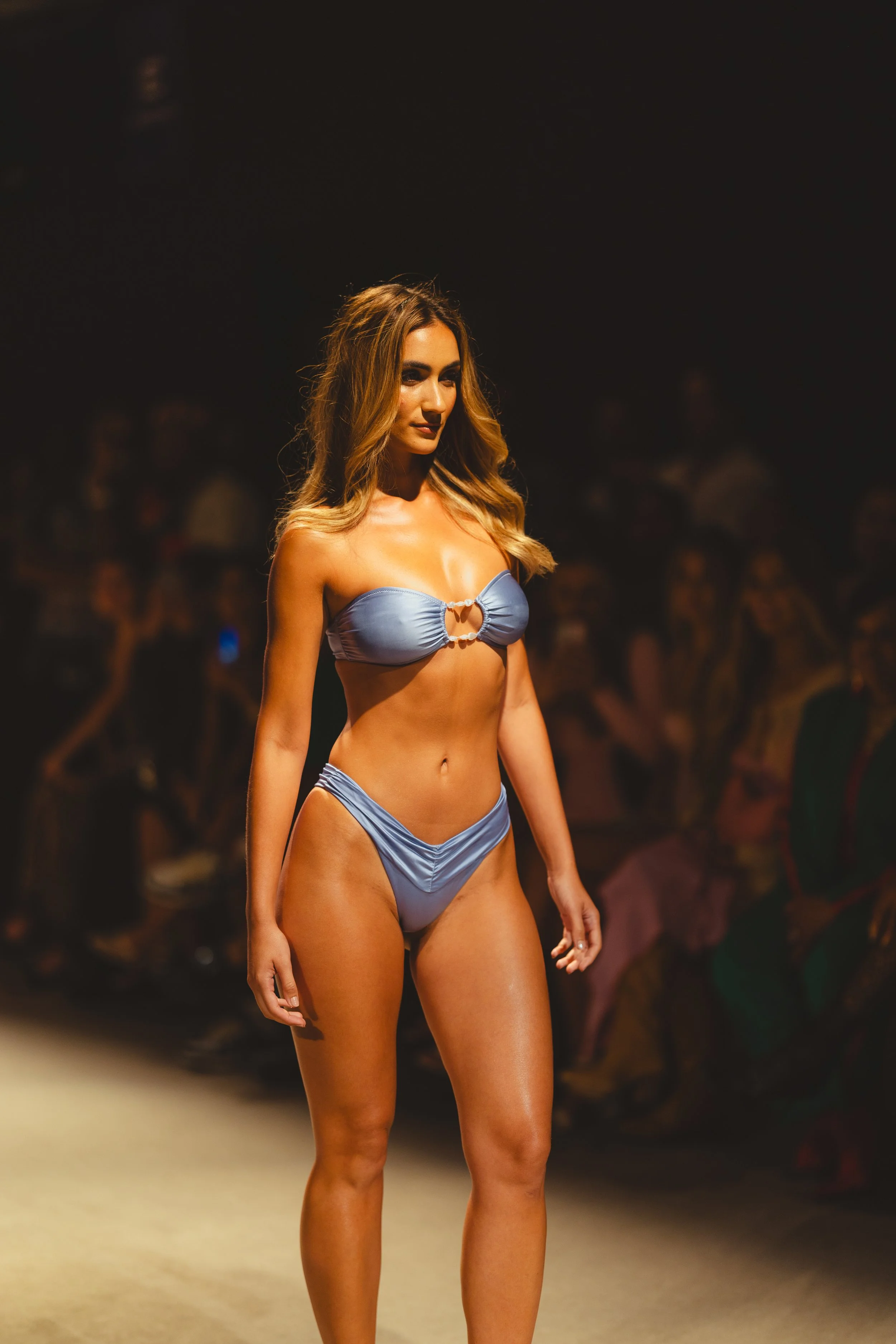A woman walking on a runway during a fashion show, wearing a light blue bikini with a ring detail on the top.
