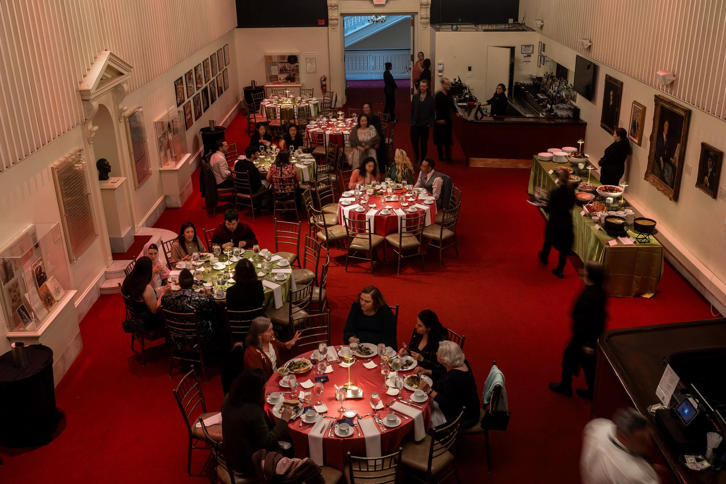 A banquet hall with round tables covered in red and green tablecloths, set for a meal with plates, glasses, and silverware. People are sitting and talking, with some standing and walking around. A bar area with bartenders and a buffet table with food
