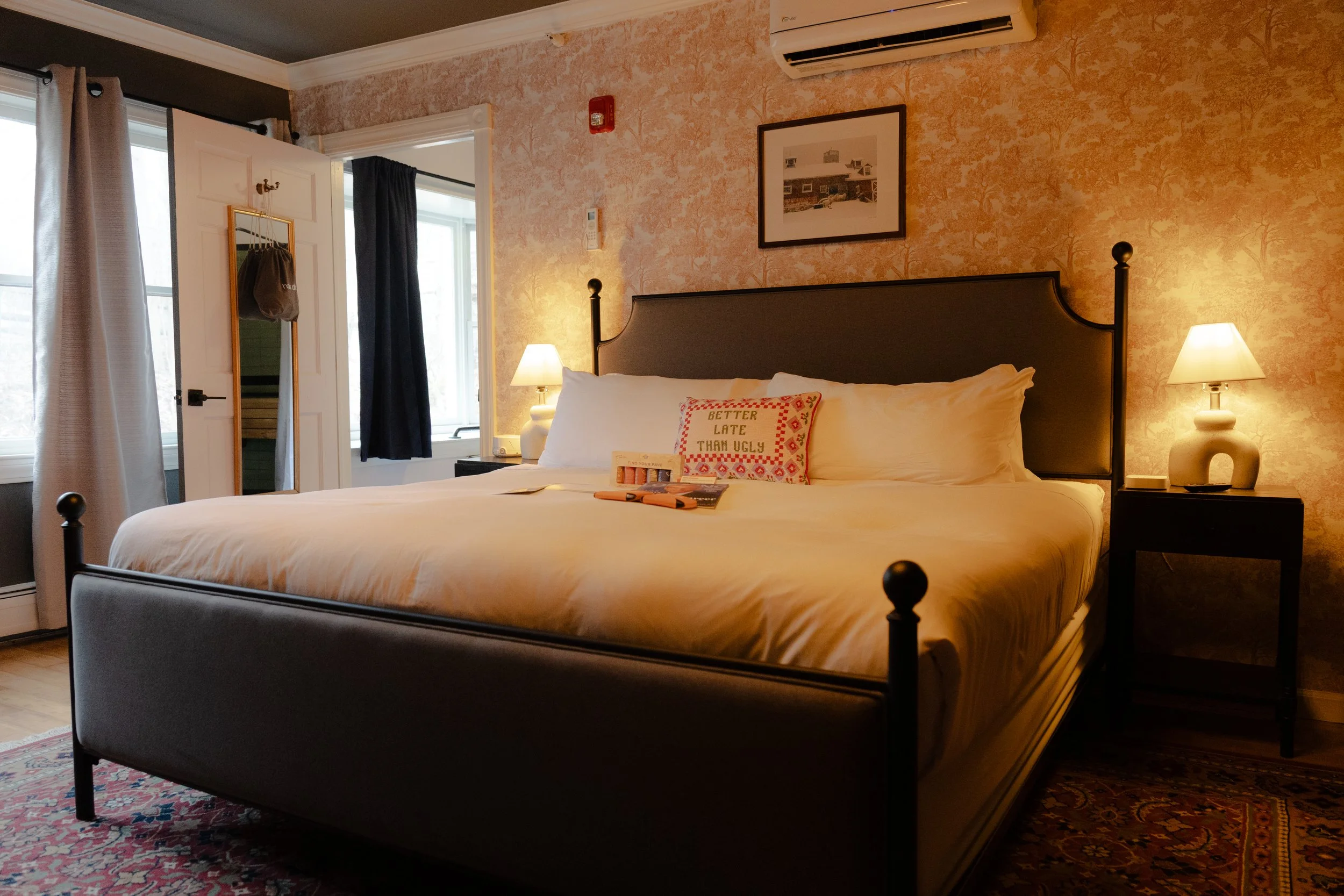 A cozy bedroom with a large bed and a dark headboard, two bedside lamps, and framed artwork on the wall. A decorative pillow with the words 'Better Late Than Ugly' is on the bed, and a mirror, window with curtains, and an air conditioning unit are vi