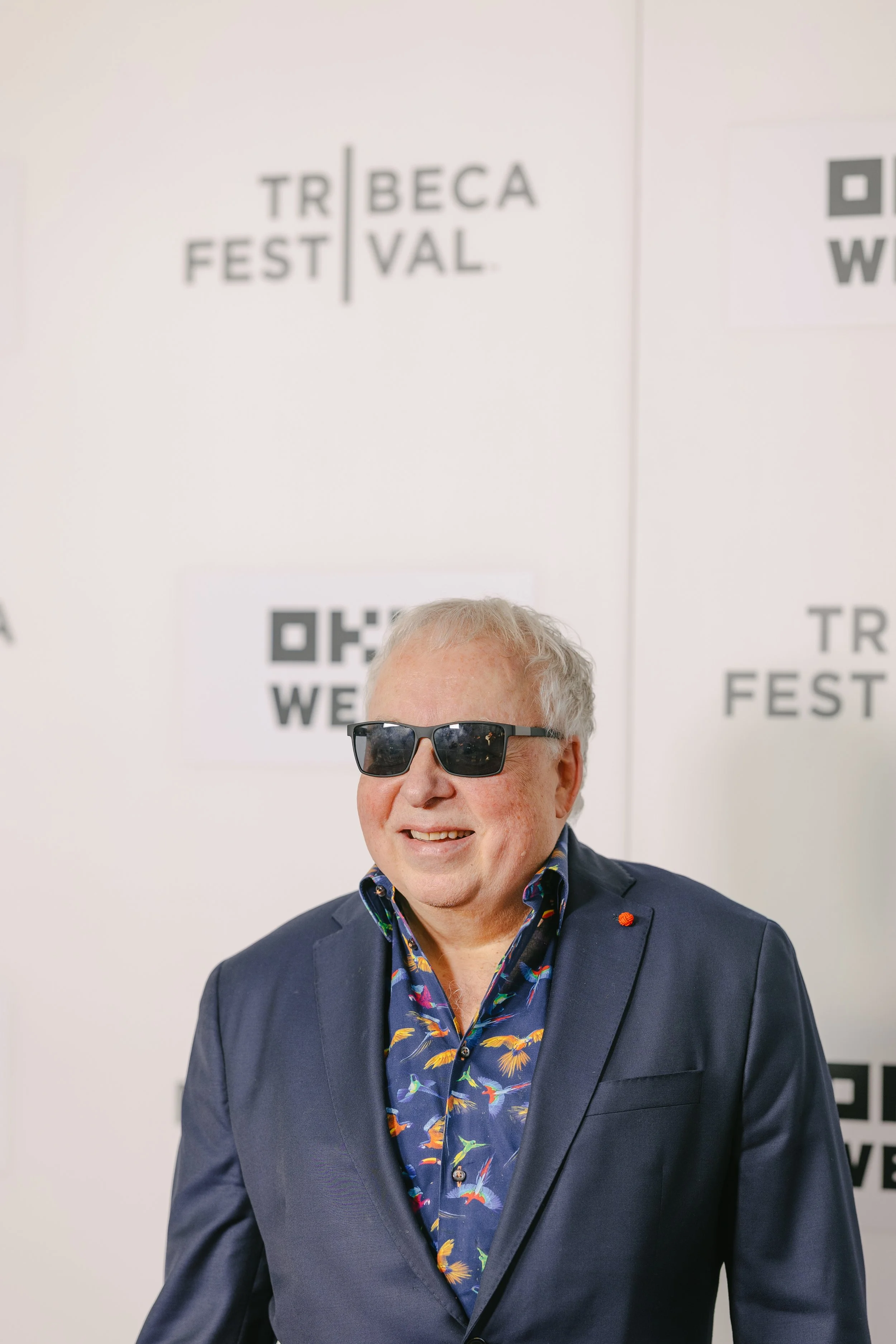 Man wearing black sunglasses, a navy blazer, and a blue shirt with a colorful bird pattern at an event with Tribeca Film Festival and WeWork signage in the background.