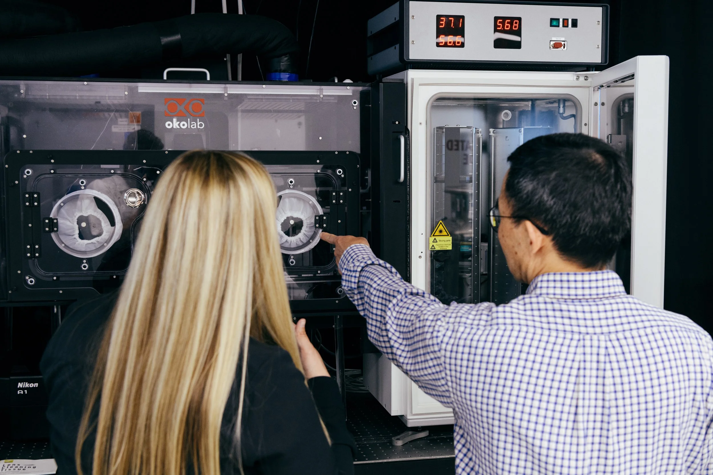 Two scientists, a woman with long blonde hair and a man with black hair, are working with a scientific instrument in a laboratory. The woman is observing and the man is pointing at the instrument which contains circular components. The laboratory equ