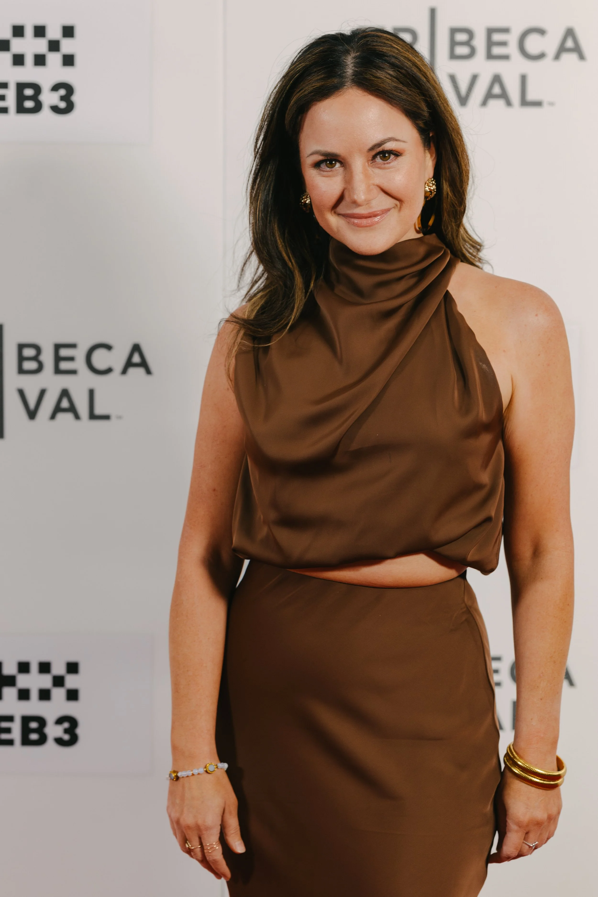 A woman in a chocolate brown satin dress with a high neckline and sleeveless design, standing in front of a white backdrop with BFI and BECA Festival logos.