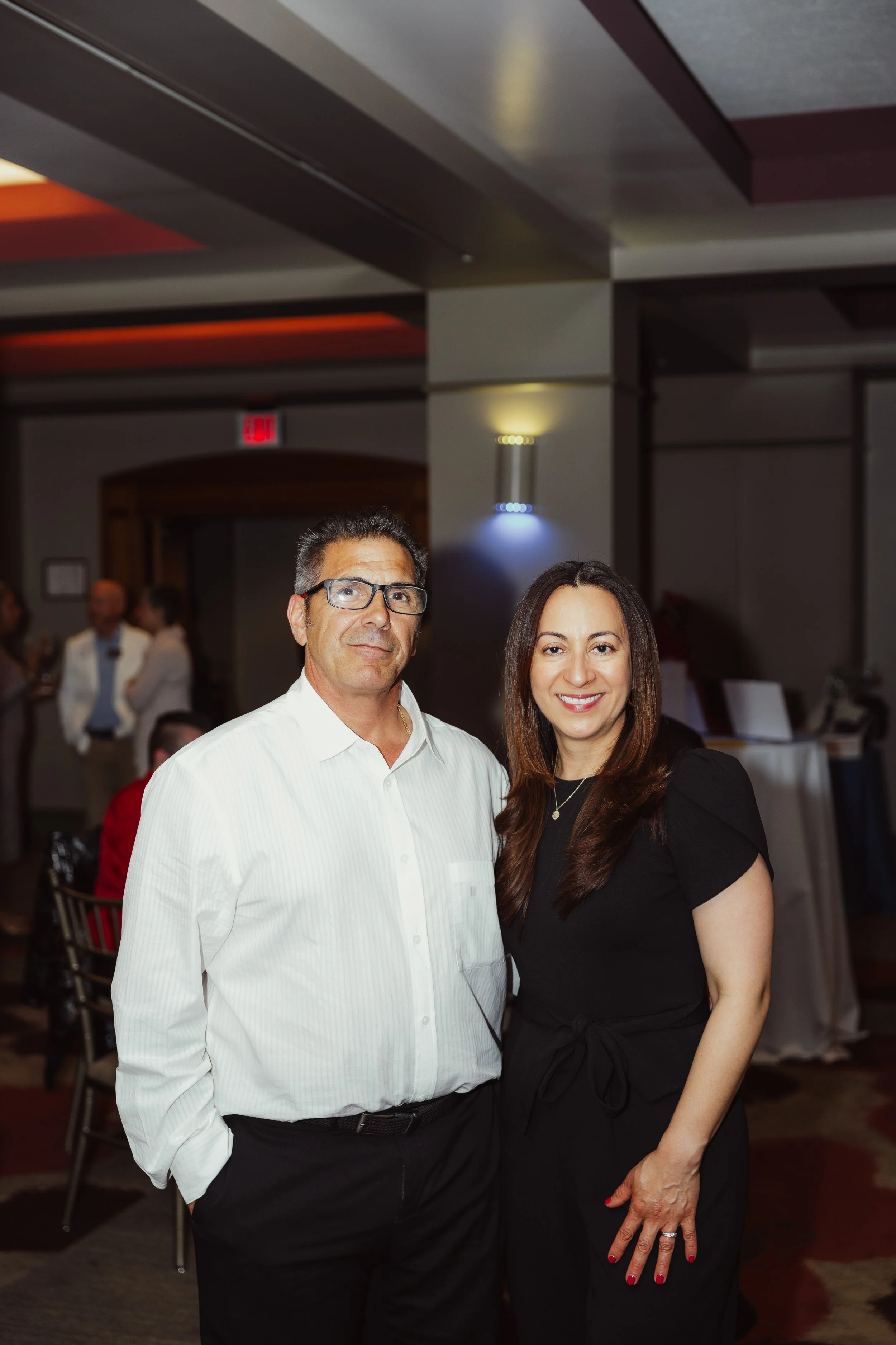 A man with glasses wearing a white shirt and a woman in a black dress with red lipstick and nail polish standing together inside a restaurant or event venue. There are other people in the background.