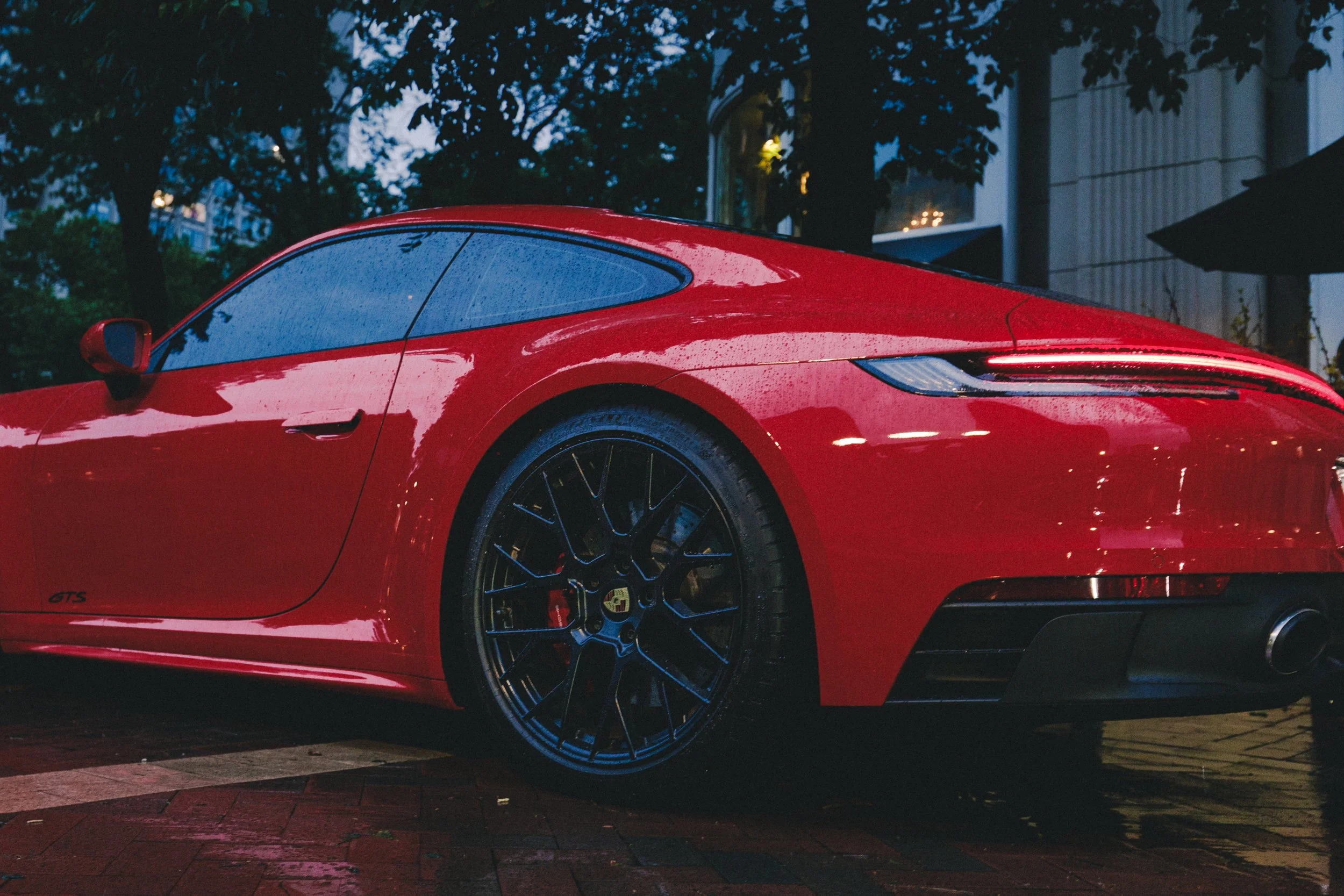 Close-up of a red sports car parked on a city street during rainy weather, with reflections of trees and buildings on its wet surface.