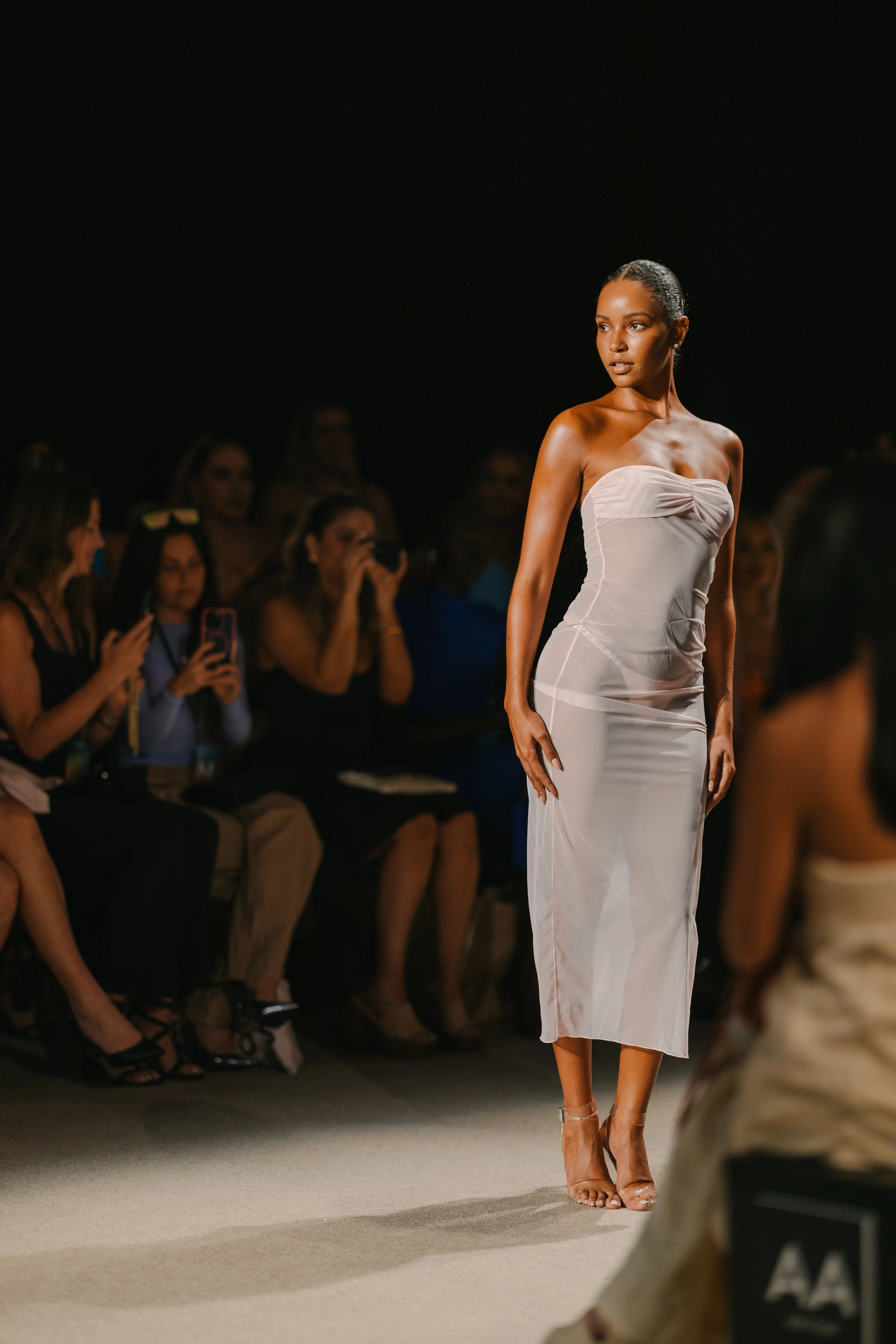A female model walking down the runway at a fashion show, wearing a strapless white dress and clear heels, with an audience seated on both sides taking photos.