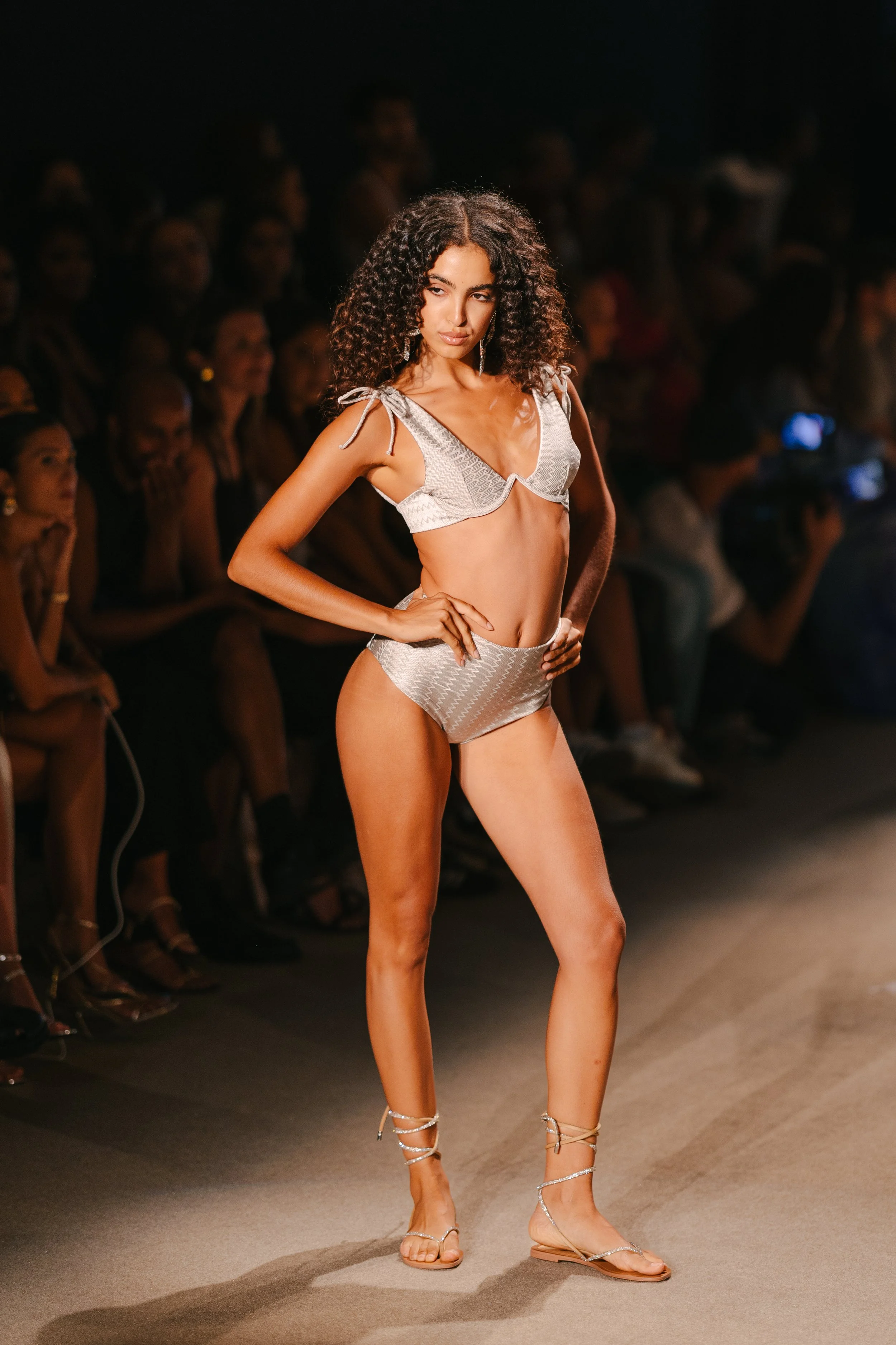 A female model walking a runway at a fashion show, wearing a silver two-piece swimsuit and silver strappy sandals, with audience members watching in the background.