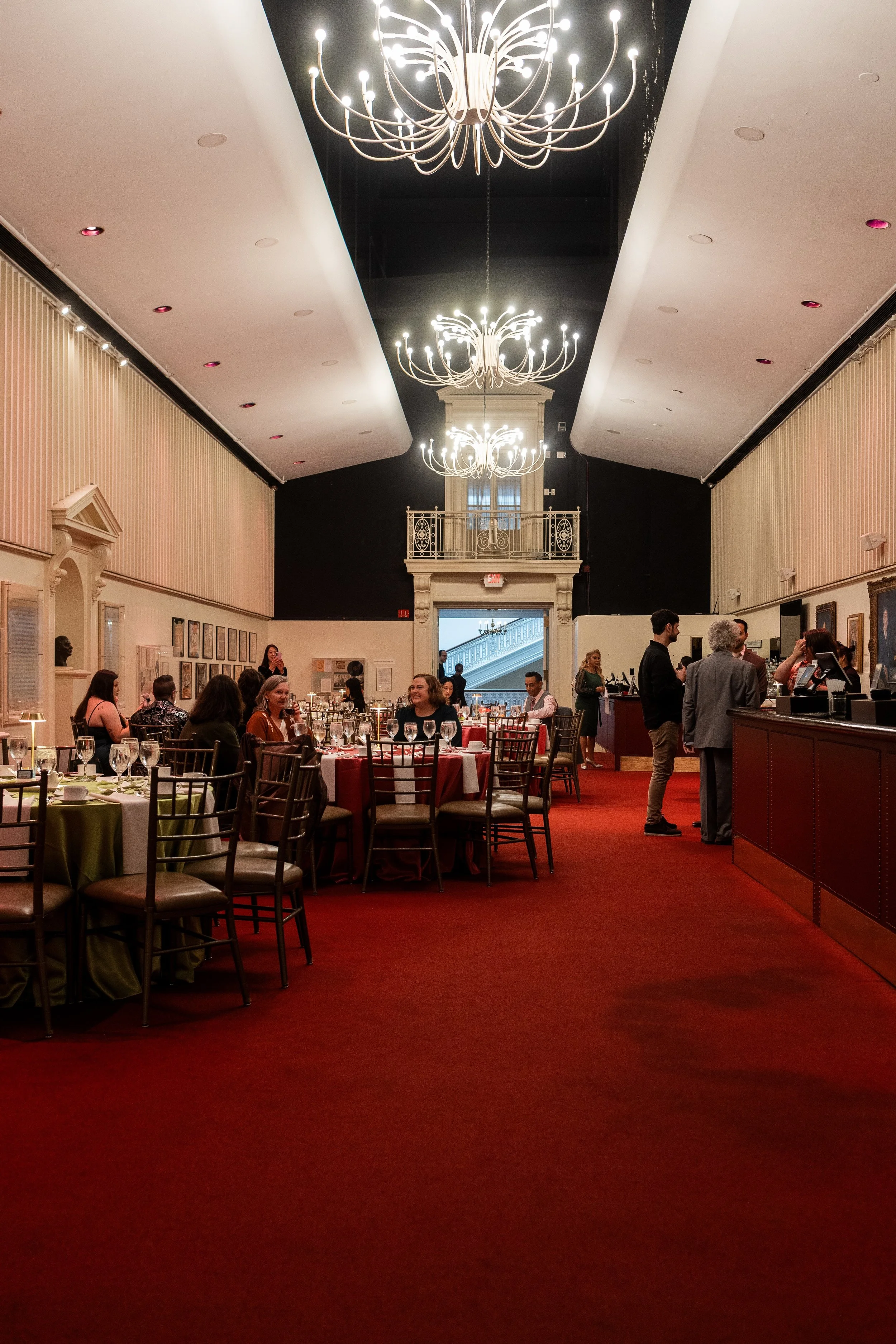 An elegant banquet hall with a high ceiling, ornate chandeliers, and a deep red carpet. Several tables are set with glasses and plates, and people are gathered, mostly seated and engaged in conversations. The far wall features framed artworks, and th