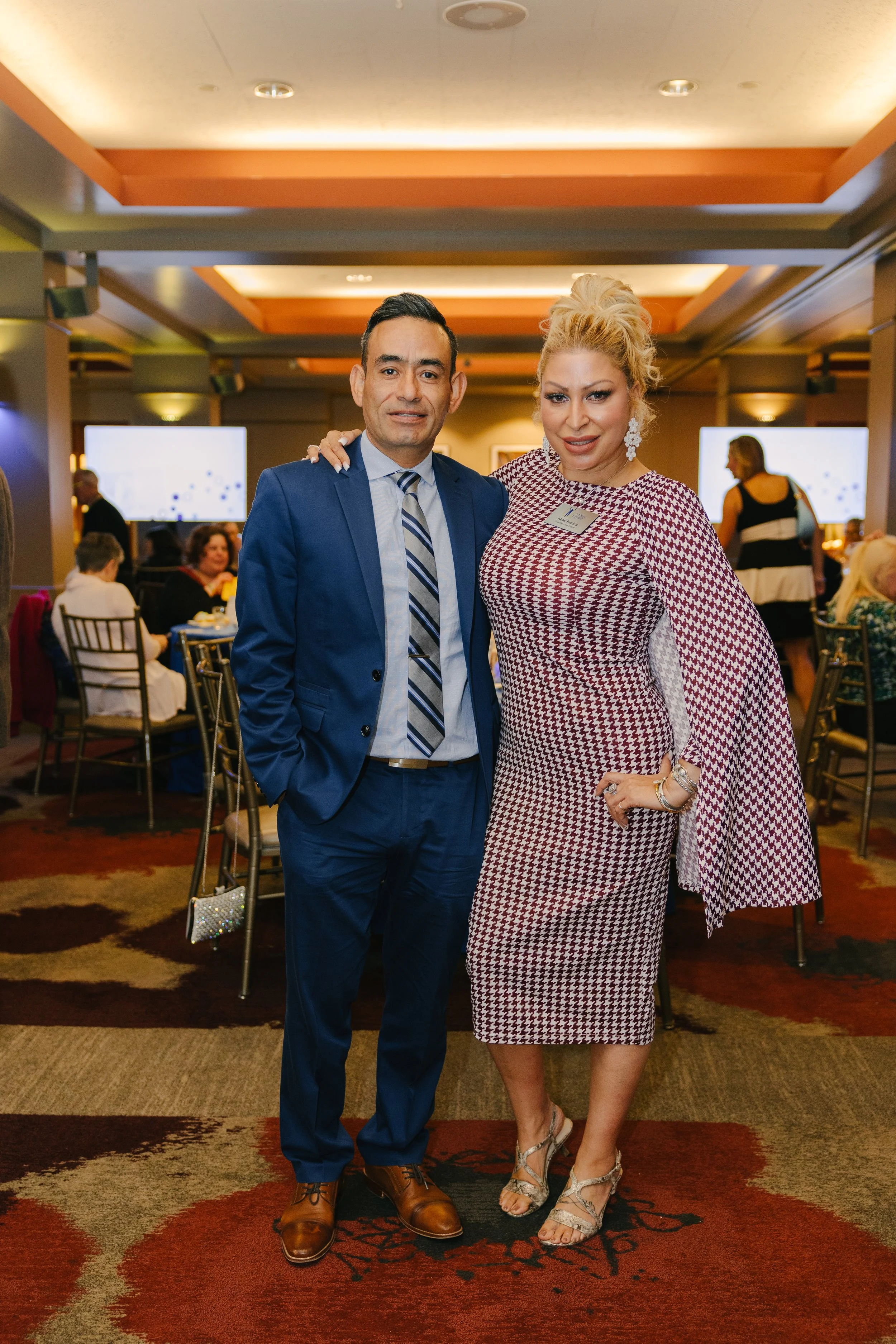 A man and a woman are standing together in a banquet hall, smiling at the camera. The man wears a blue suit with a striped tie and brown shoes. The woman wears a red and white patterned dress, large earrings, and high heels. There are other people se