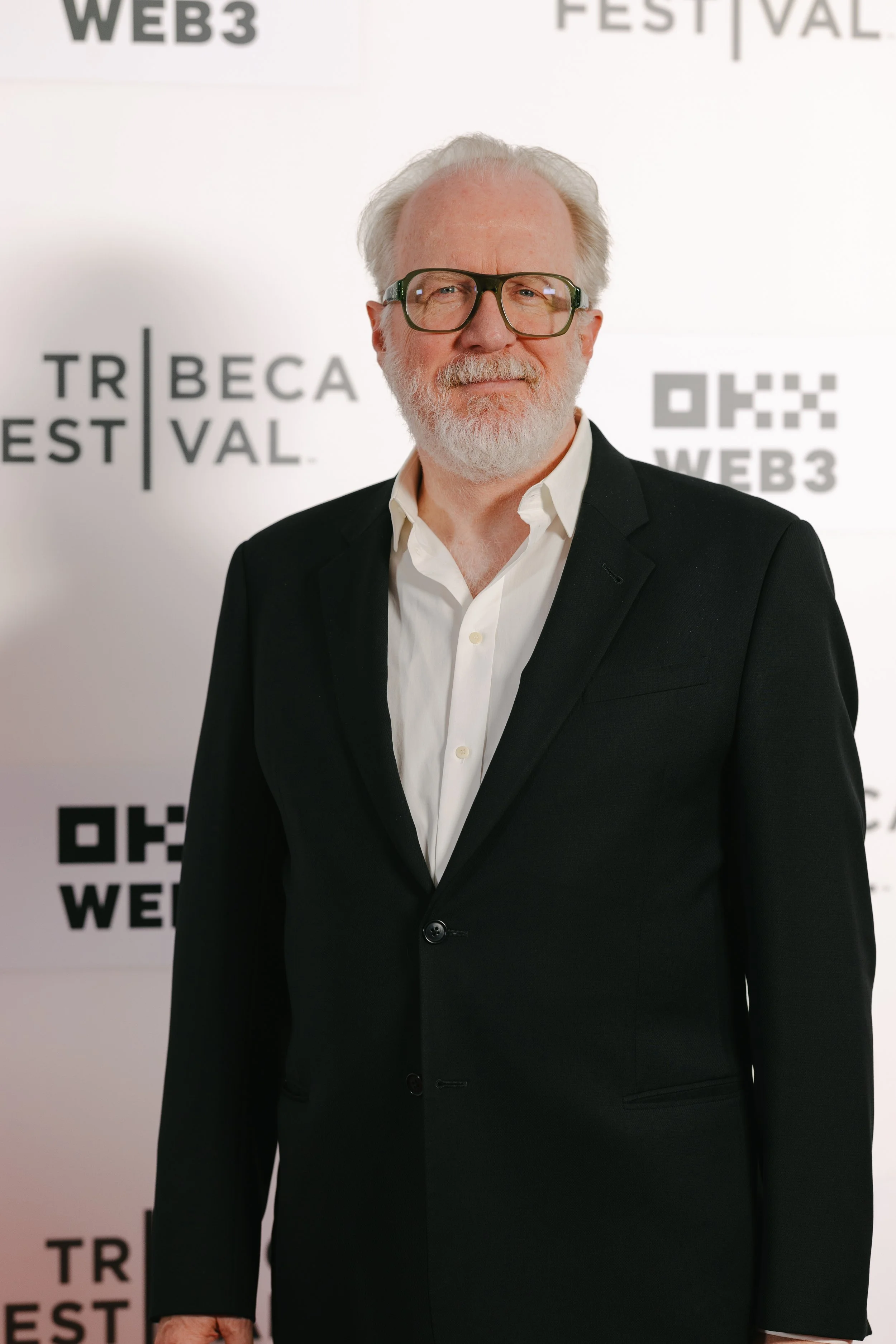 An older man with white hair and beard, glasses, wearing a black suit and white shirt, standing in front of a backdrop with logos and text related to Web3 and Tribeca Festival.