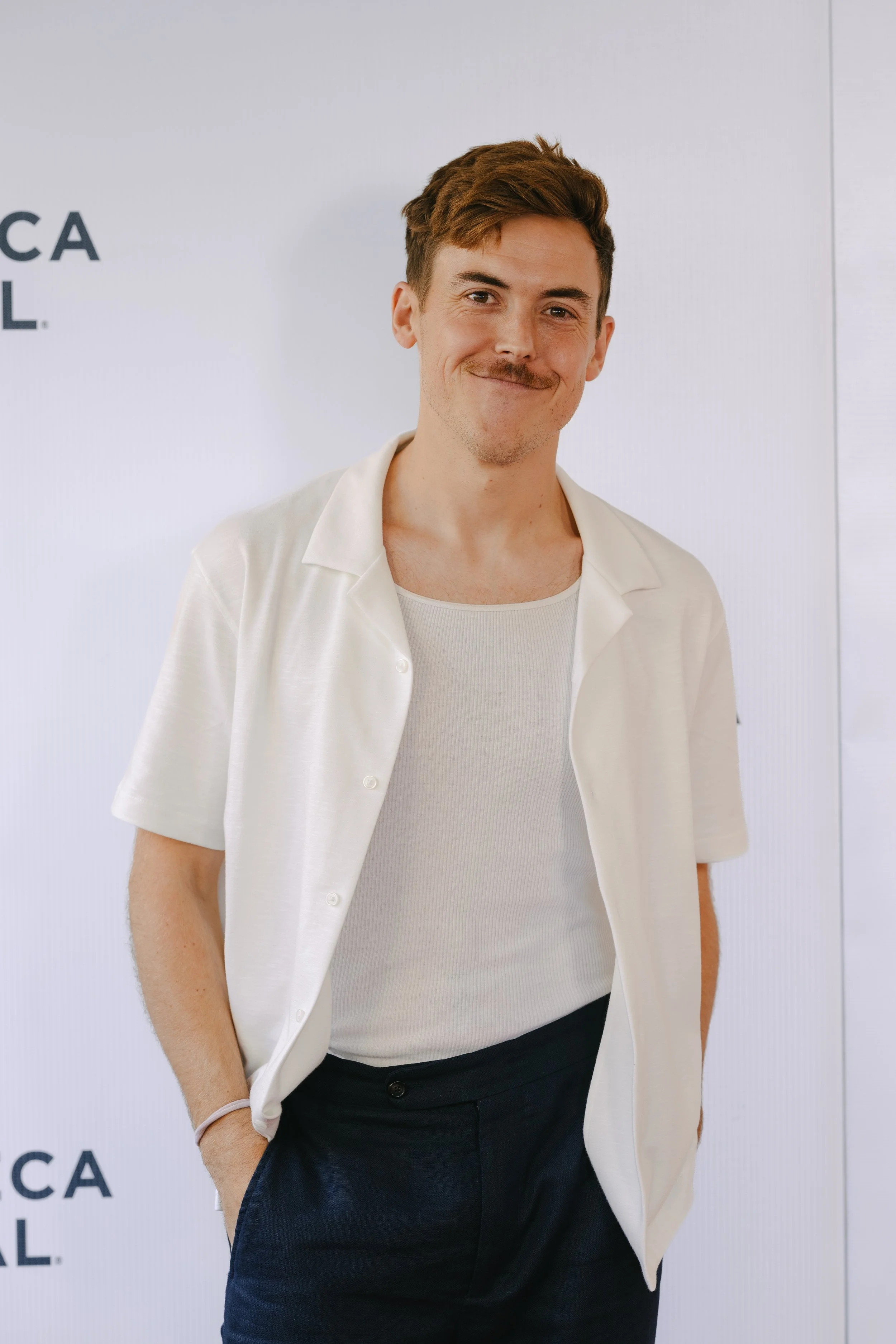 A young man with brown hair and a mustache, wearing a white short-sleeved shirt over a white tank top, standing against a white background with partial text visible.