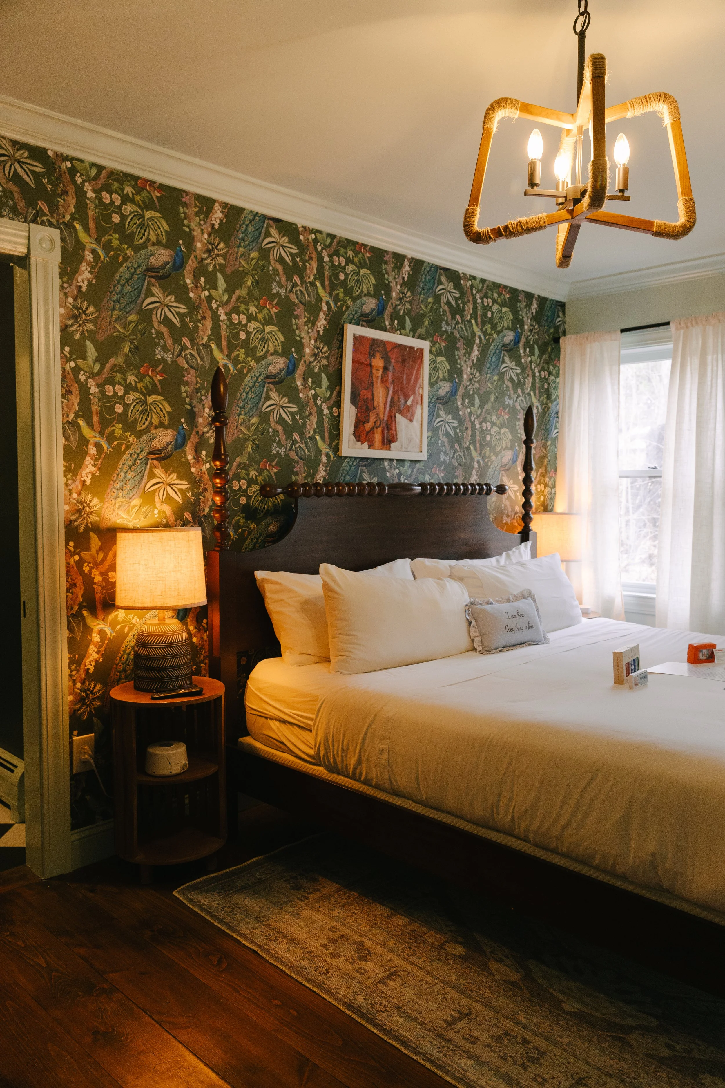 A bedroom with a dark wood bed frame, white bedding, patterned wallpaper with birds and flowers, two lamps on side tables, a framed picture above the headboard, a window with white curtains, and a modern ceiling light fixture.