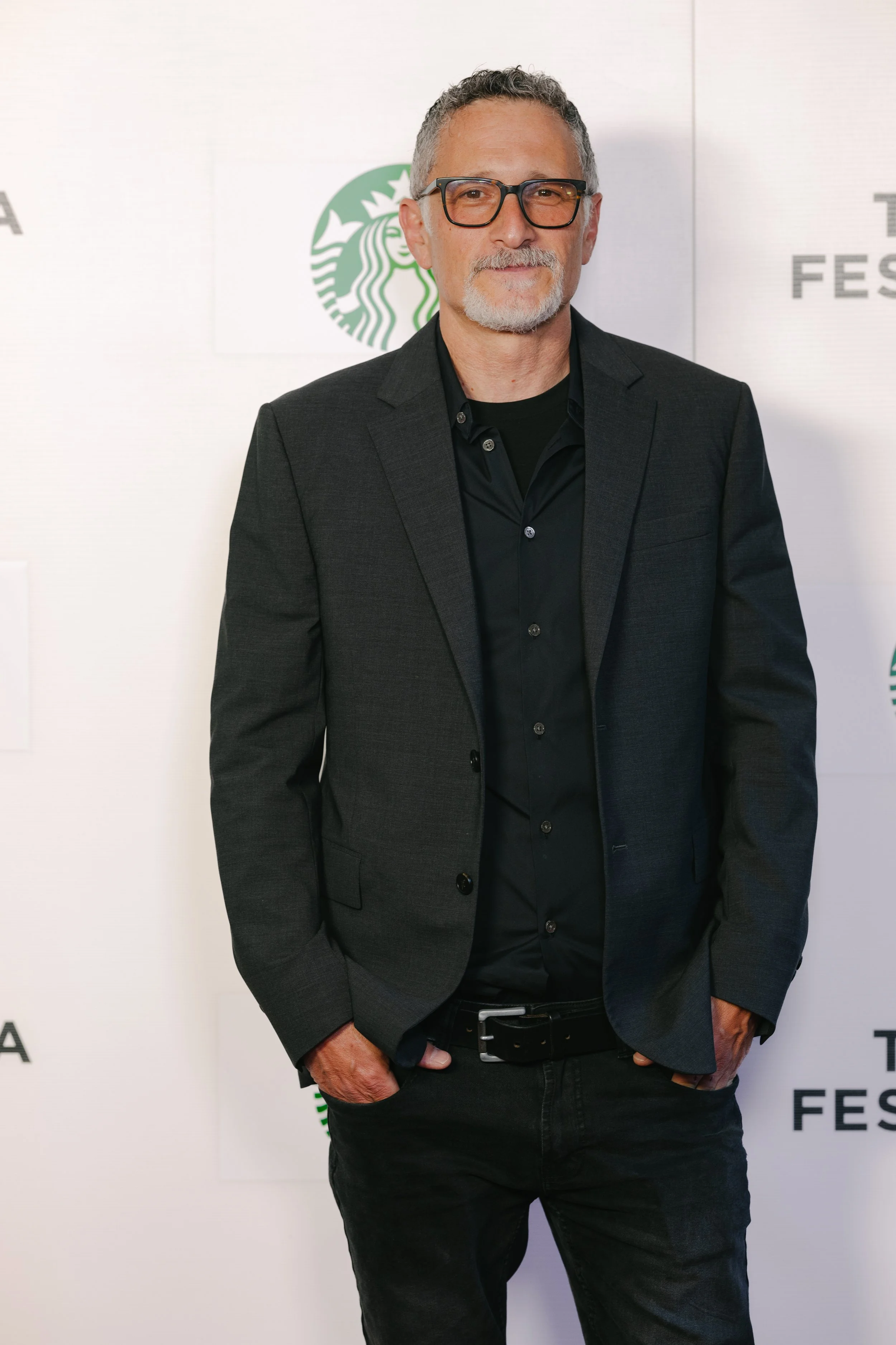 A middle-aged man with gray hair and beard wearing glasses, a dark blazer, and black pants, standing in front of a white background with Starbucks logos.
