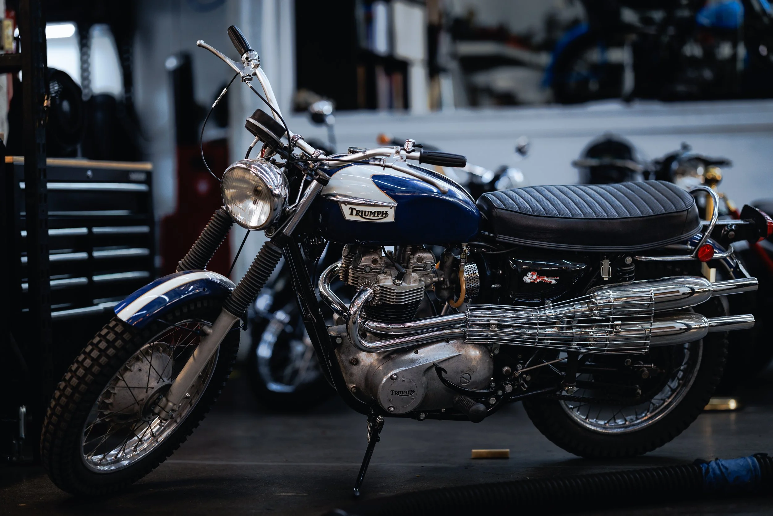 A vintage blue and white Triumph motorcycle parked in a garage with tools and other motorcycles in the background.