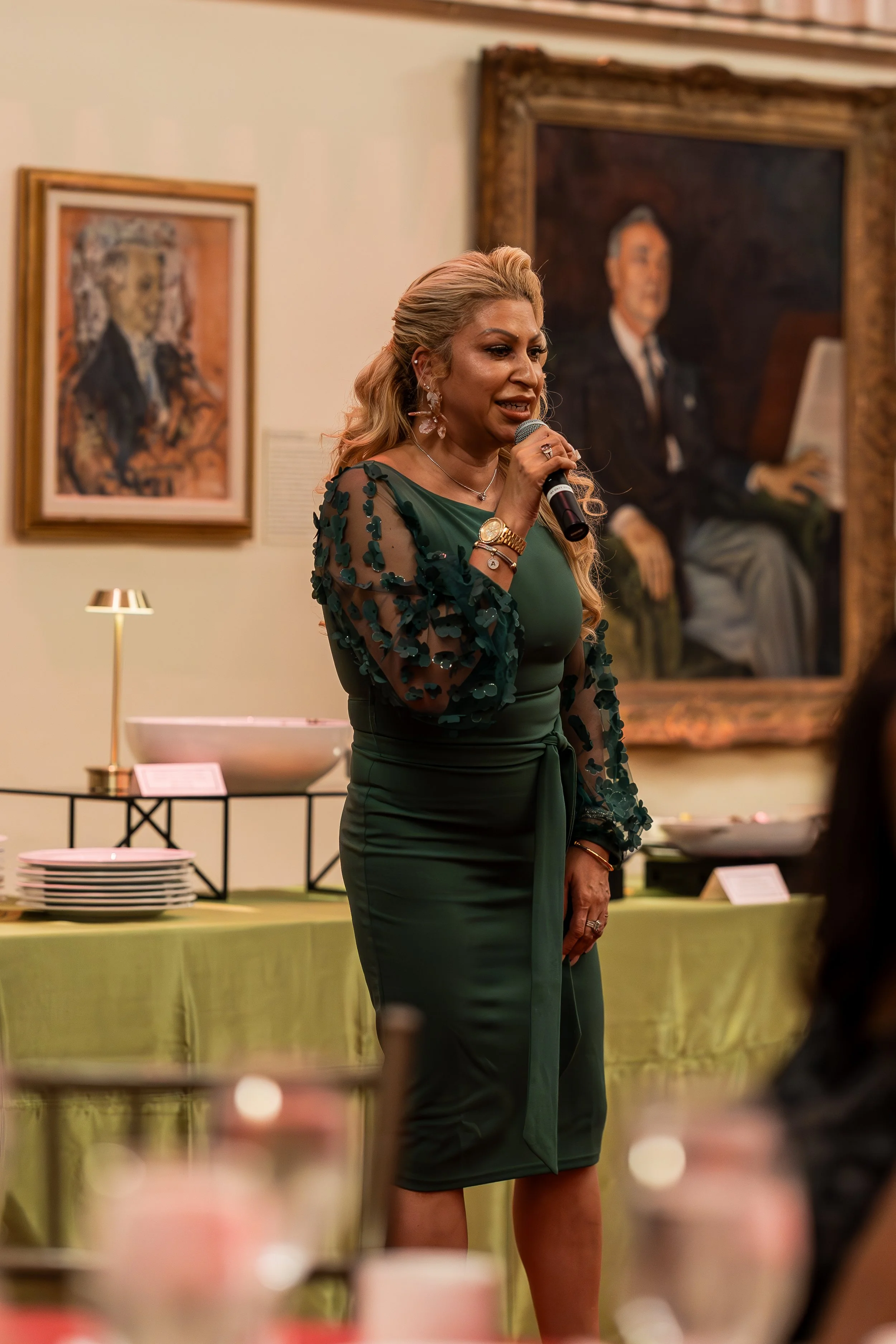 A woman with blonde hair in a green dress and floral sheer sleeves is speaking into a microphone at an indoor event. Behind her, there are framed paintings on the wall and a table with plates and serving dishes.