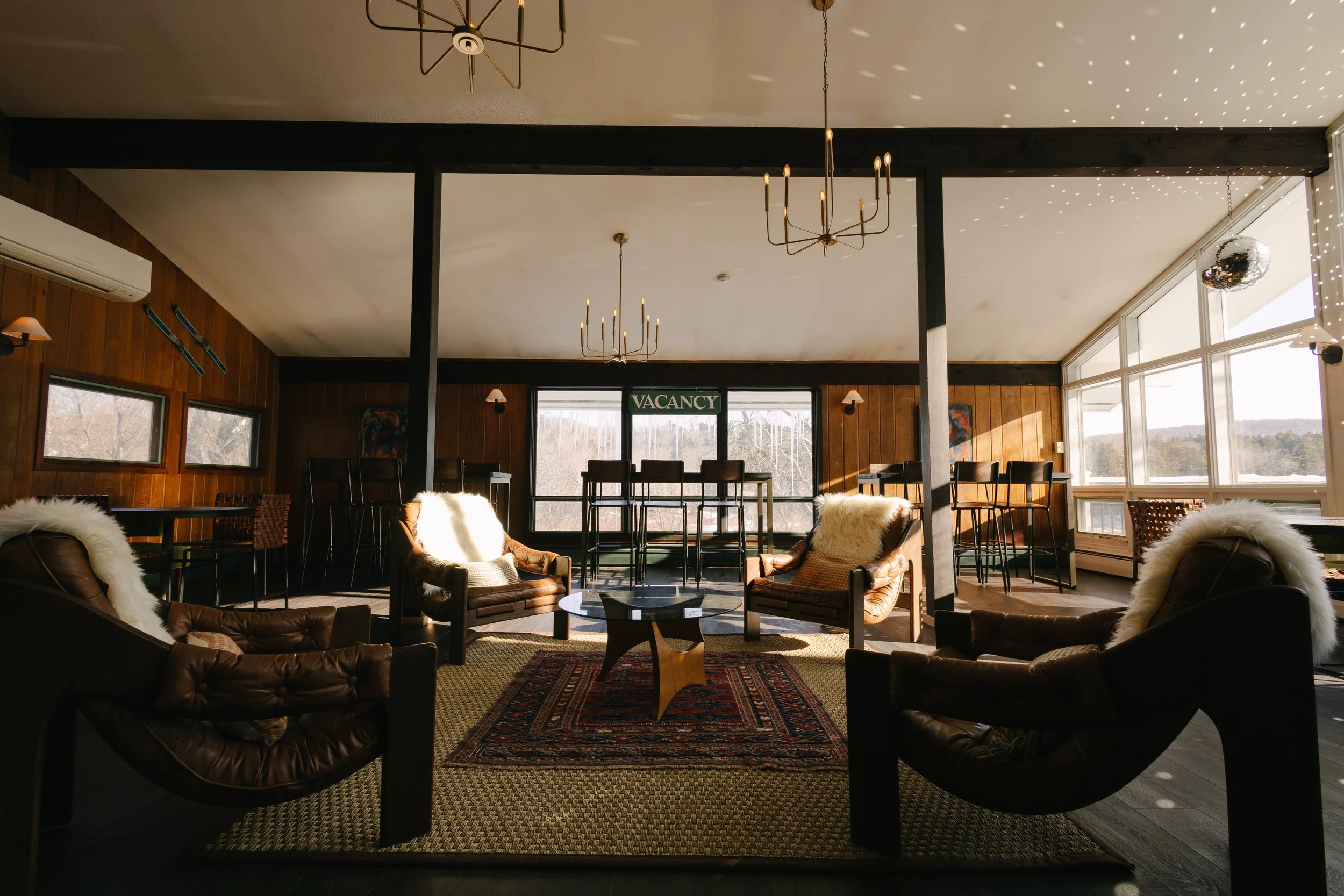 A cozy lounge with wooden walls, leather armchairs with fur throws, a patterned rug, large windows letting in natural light, and barstools near the window with a 'Vacancy' sign overhead.