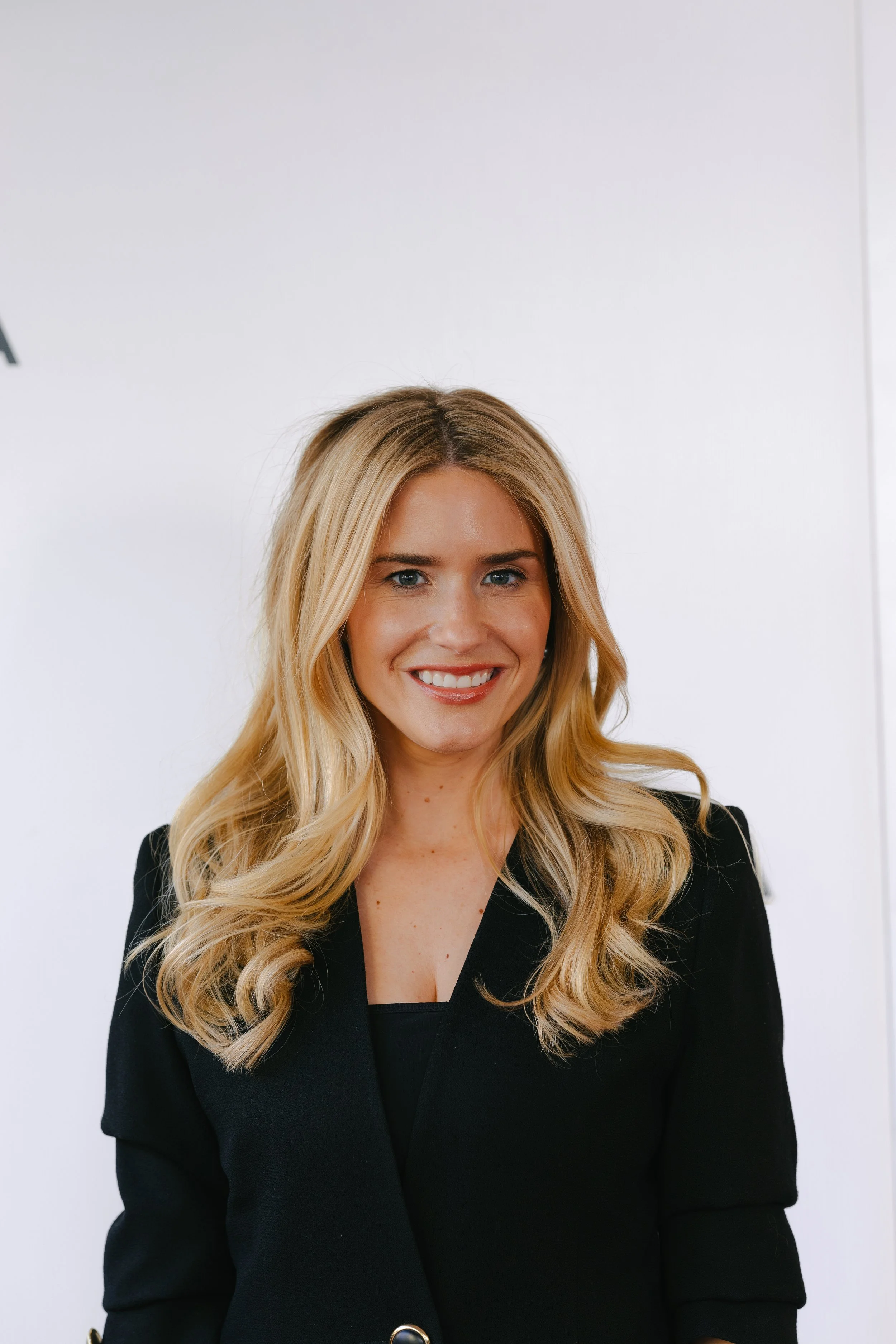 A smiling woman with long blonde hair wearing a black blazer standing against a white wall.