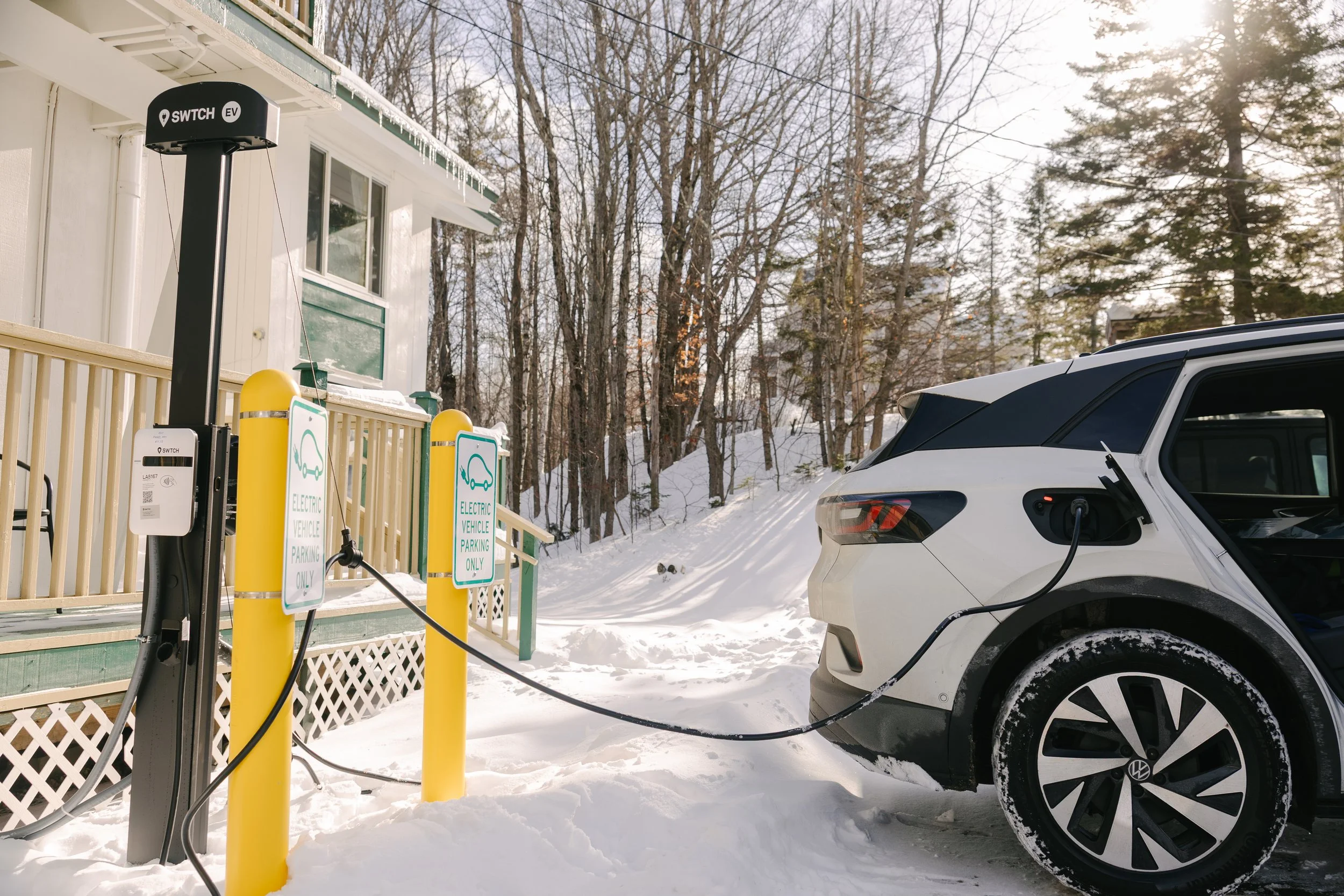 An electric vehicle charging station with a white Volkswagen ID4 plugged in, on a snow-covered driveway, with a house and trees in the background.