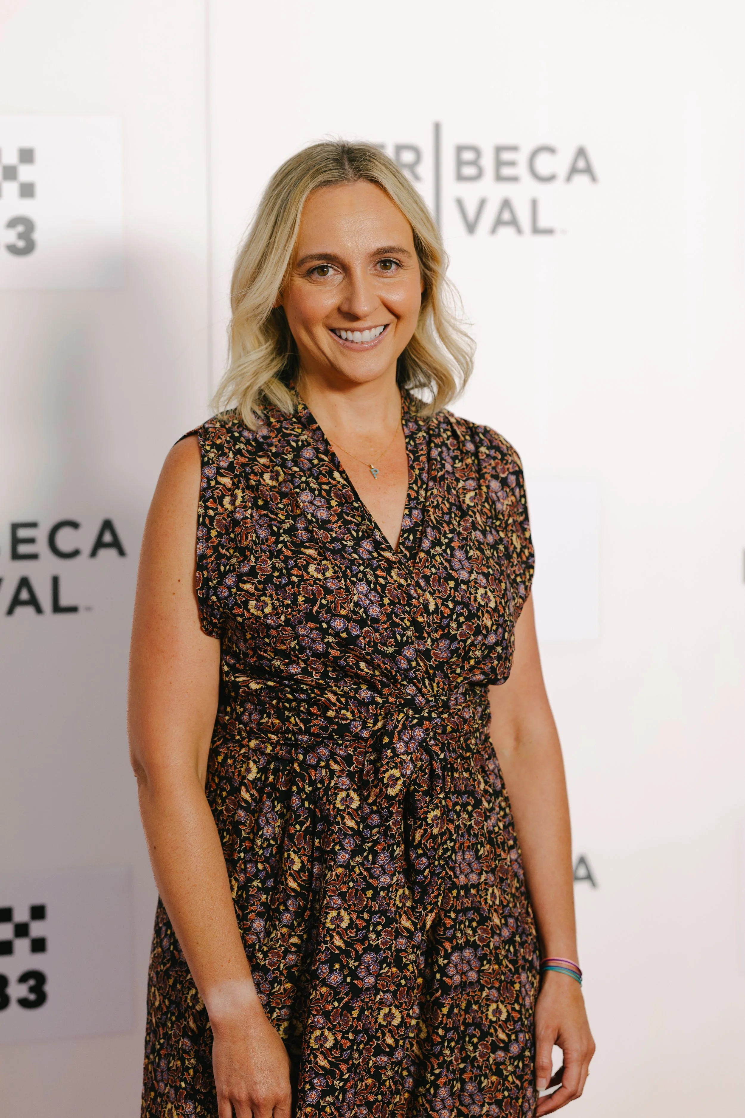 A woman with shoulder-length blonde hair standing in front of a backdrop with the words "Tribeca Film Festival." She is smiling and wearing a sleeveless, floral-patterned dress with a tie at the waist, and a necklace with a small pendant.