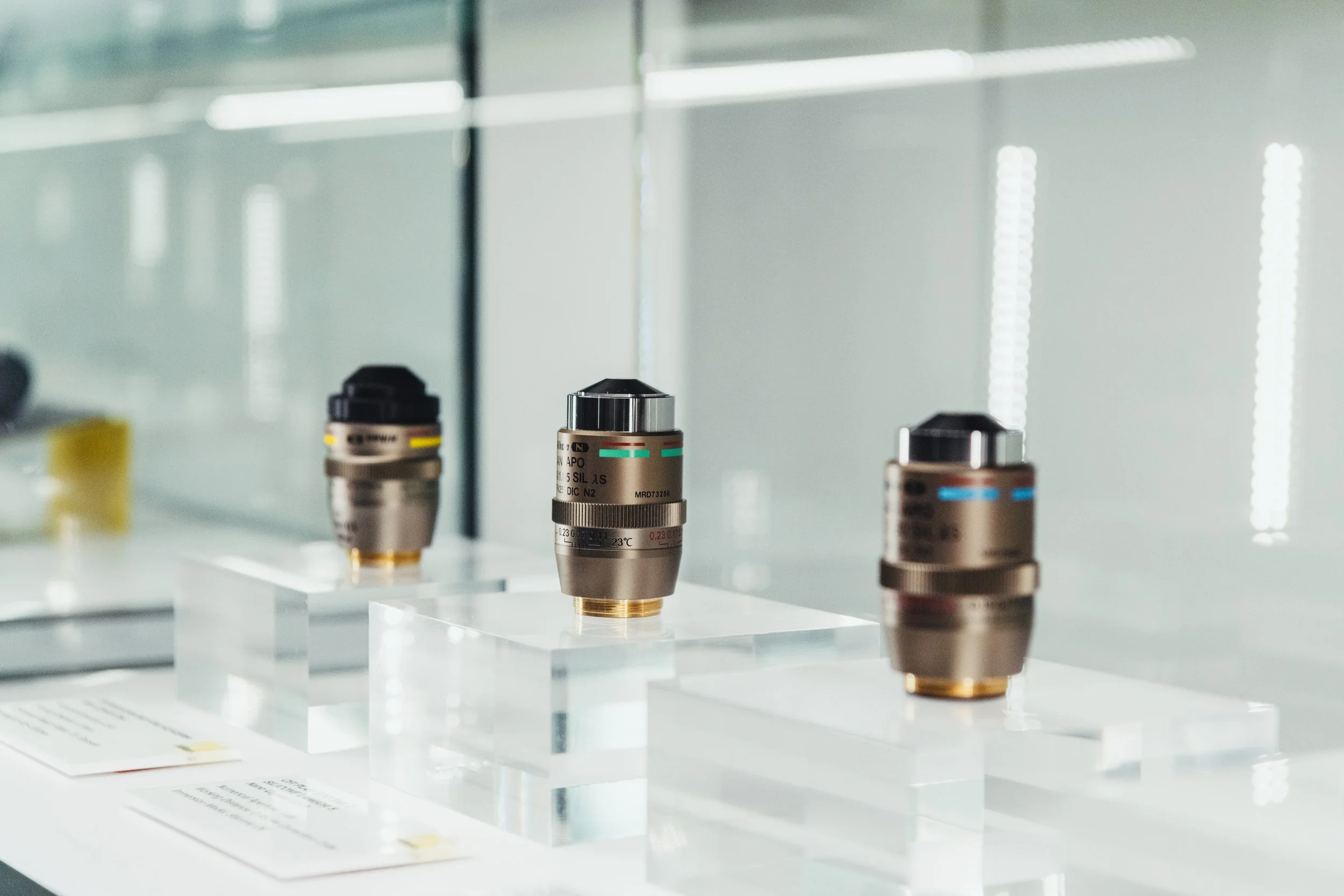 Three metal industrial camera lenses displayed on clear acrylic stands in a modern, well-lit showroom.