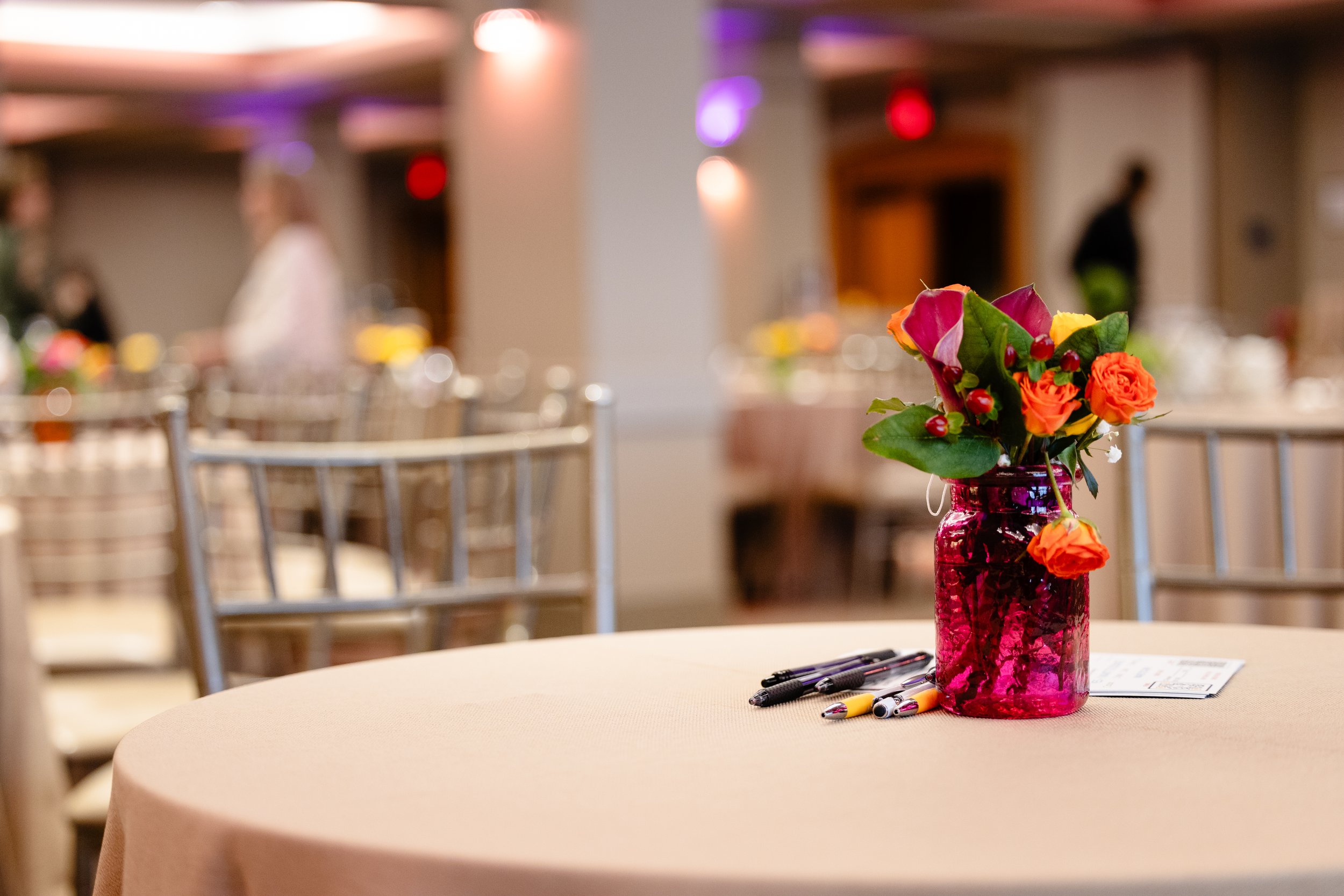 A table with a pink vase holding a bouquet of orange, pink, and yellow flowers, surrounded by pens and papers, in a softly lit conference or event space.