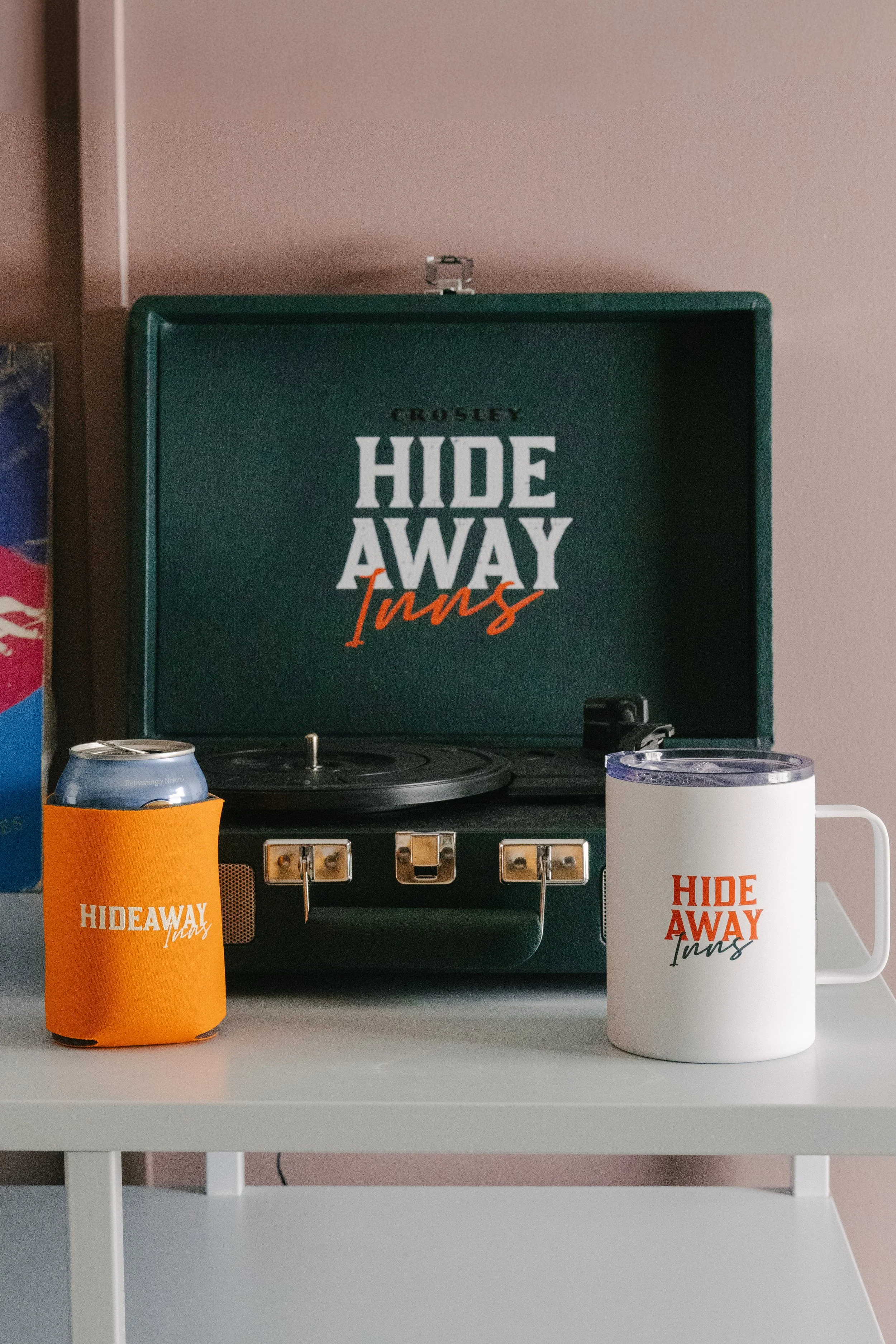 A vintage record player with a green case and the words 'Crosley Hide Away Turns' inside the lid. Two beverage containers, one orange and one white with a lid and a handle, both featuring the same logo and text, are placed on a white table in front o