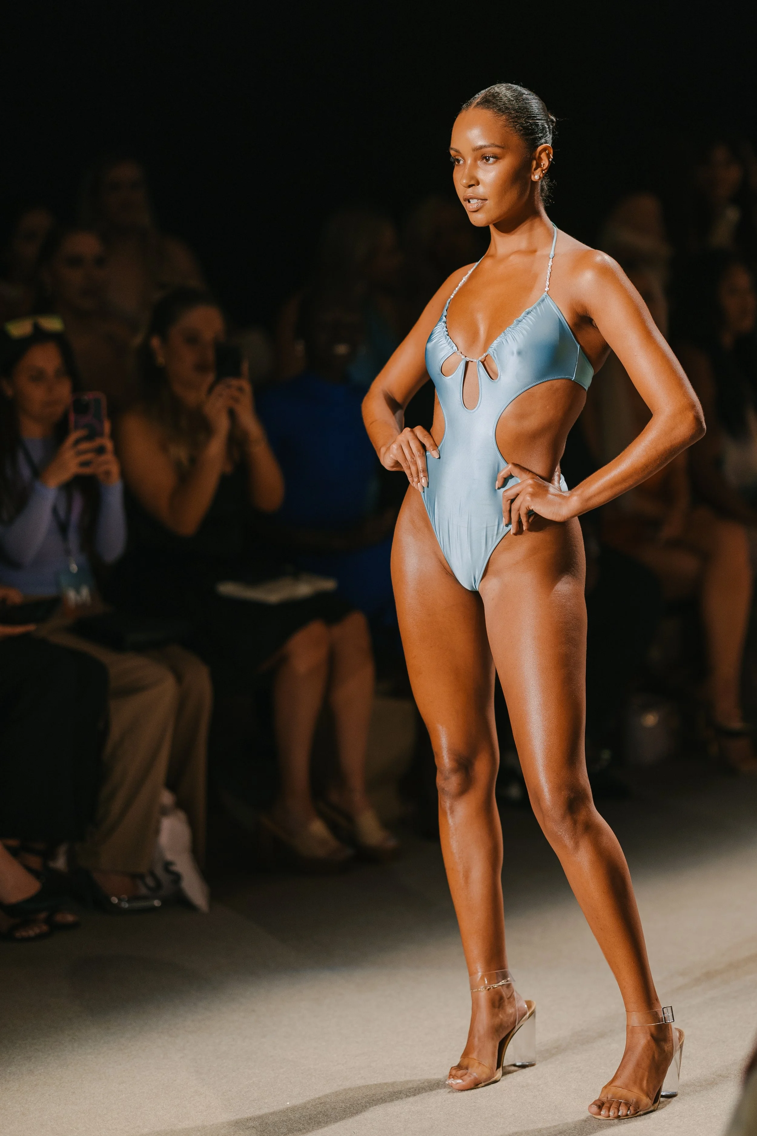 A woman modeling a light blue swimsuit on a runway at a fashion show, with an audience watching and some taking photos.