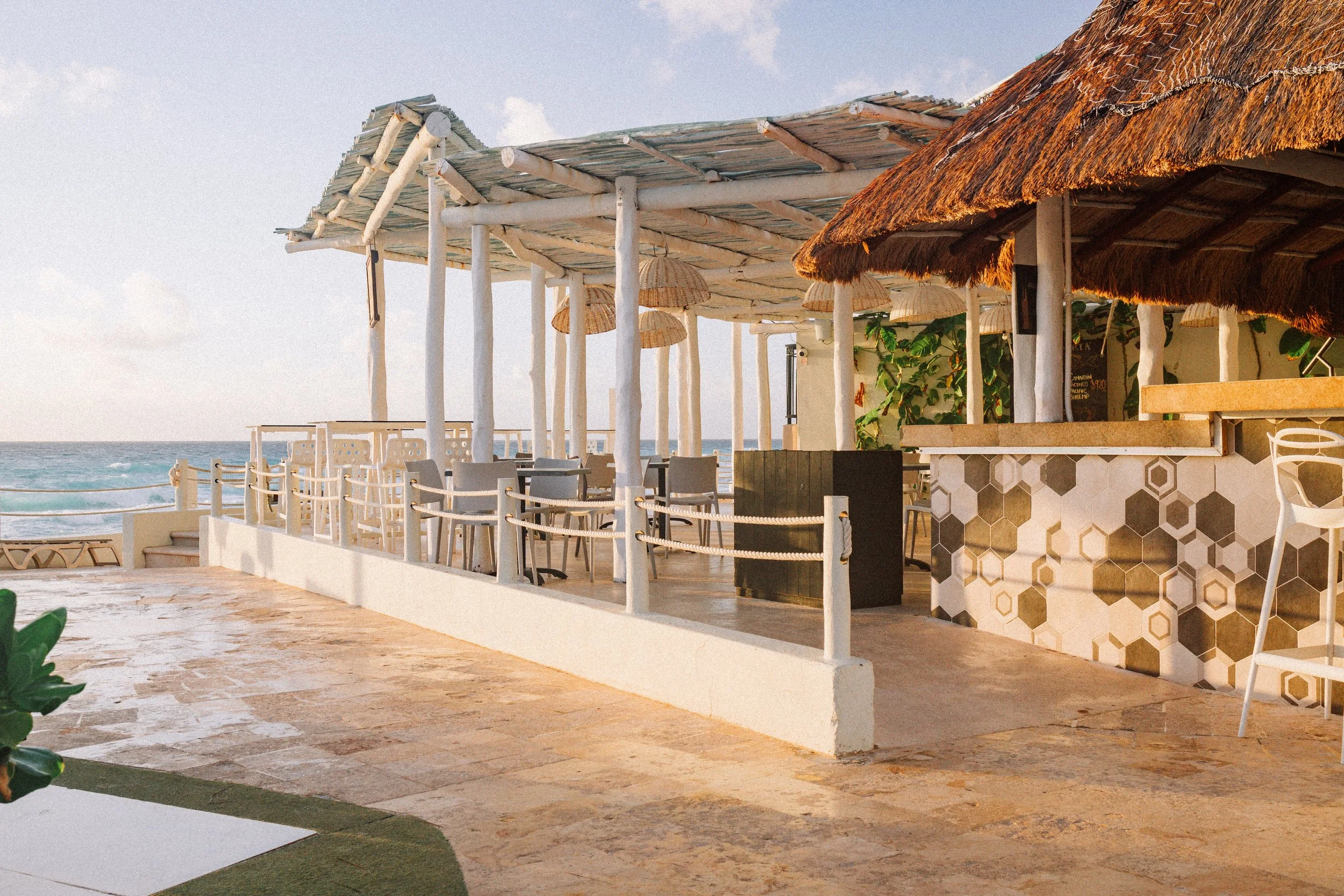Beachside outdoor restaurant with wooden tables and chairs, hanging wicker lamps, thatched roof sections, and ocean view in the background.