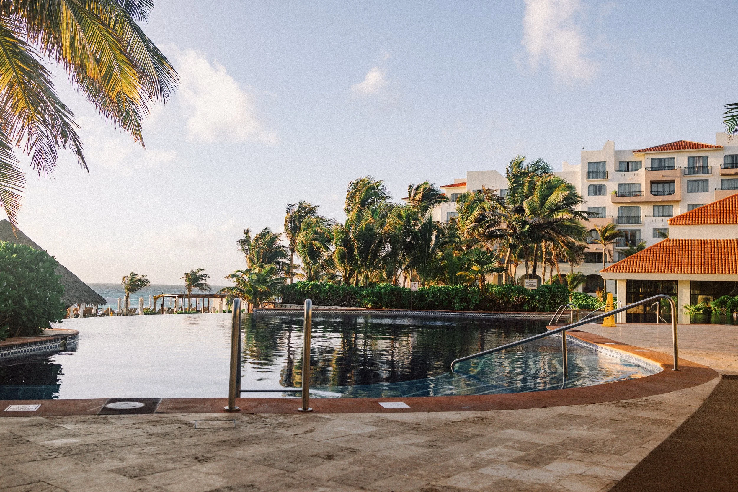 Swimming pool surrounded by palm trees and resort buildings with red-tiled roofs, ocean in background, sunny sky with some clouds.