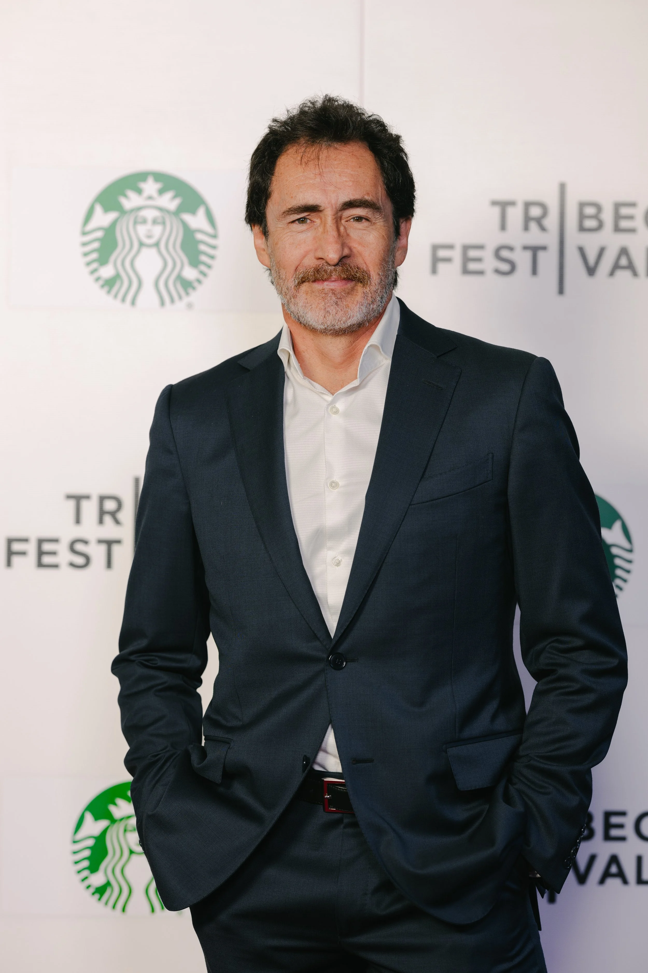 A middle-aged man with dark hair and a beard, wearing a navy blue blazer and white dress shirt, standing with hands in pockets at a film festival event.