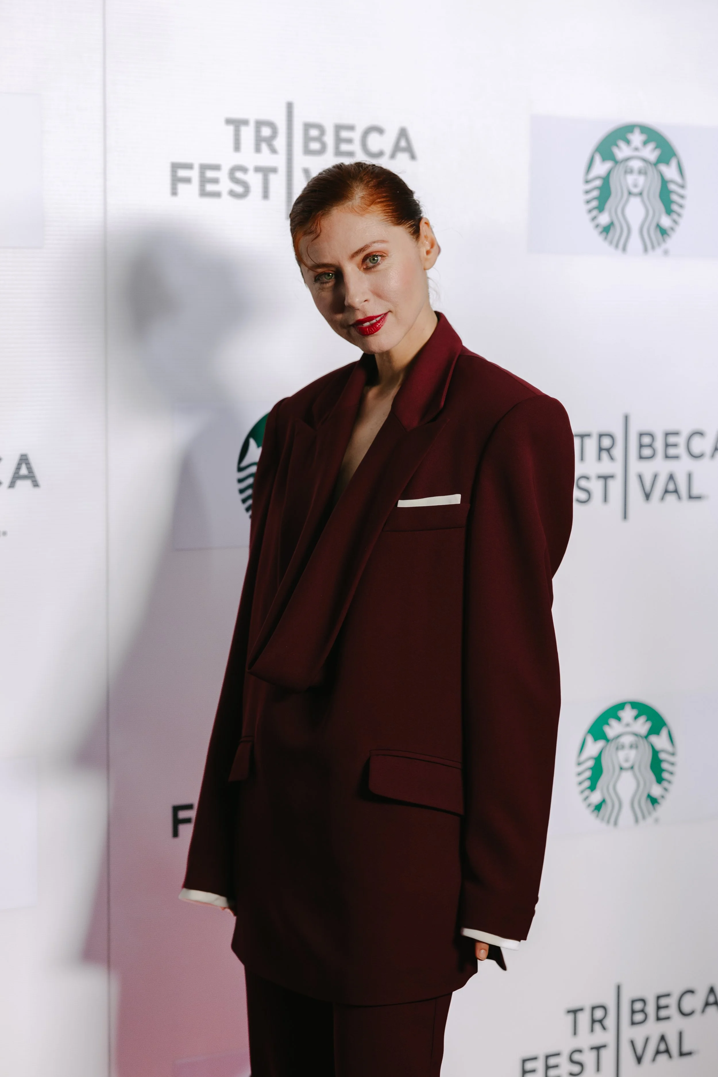 A woman in a dark red suit standing in front of a white background with logos for the Tribeca Film Festival and Starbucks.