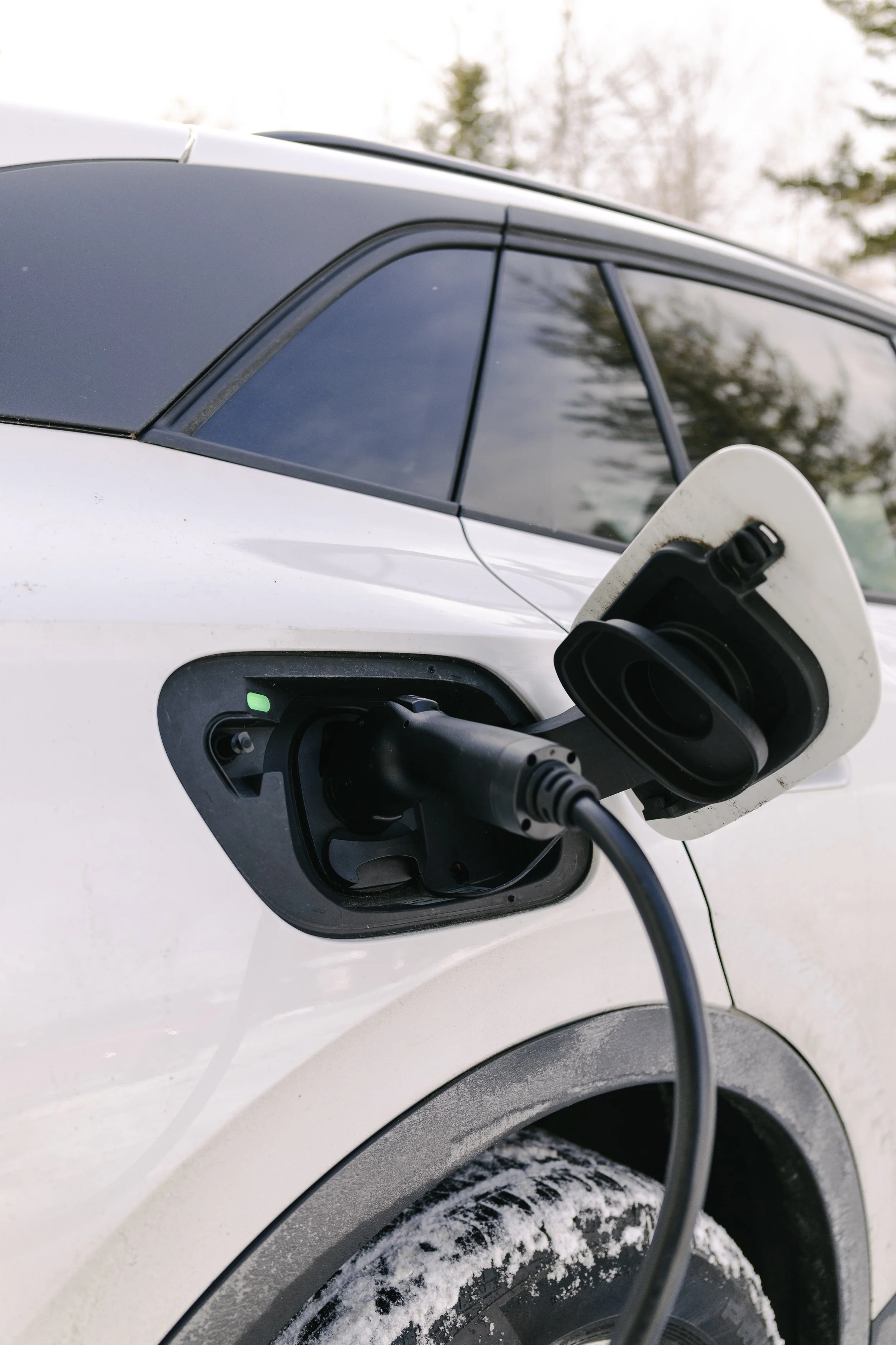 Electric vehicle charging port with charger plugged in, white car outside in winter.