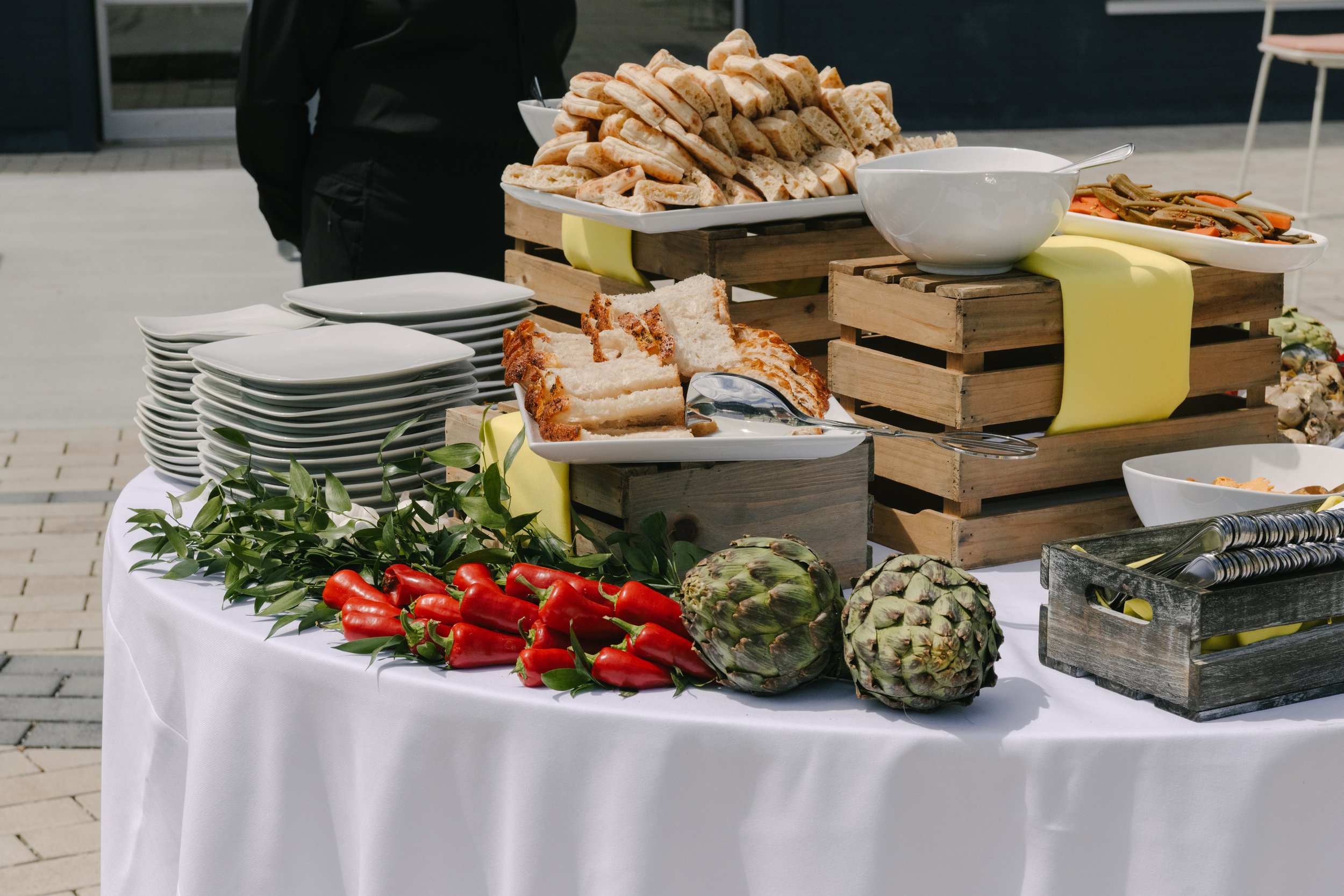 Buffet table with sliced bread, cake, artichokes, red peppers, salads, and plates, outside on a sunny day.