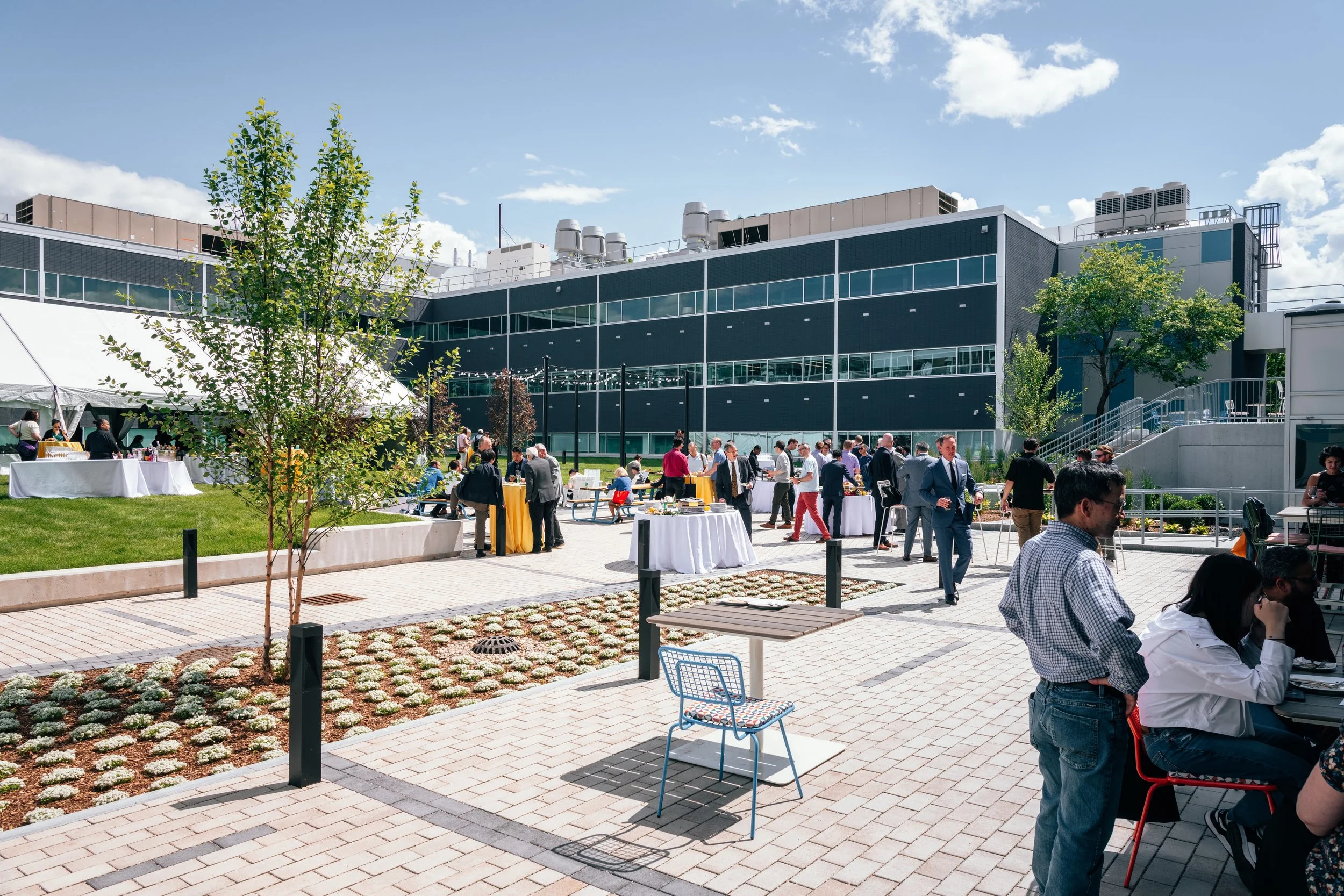 Outdoor corporate event with people socializing, buffet tables, and chairs in a paved courtyard next to a modern building under a partly cloudy sky.