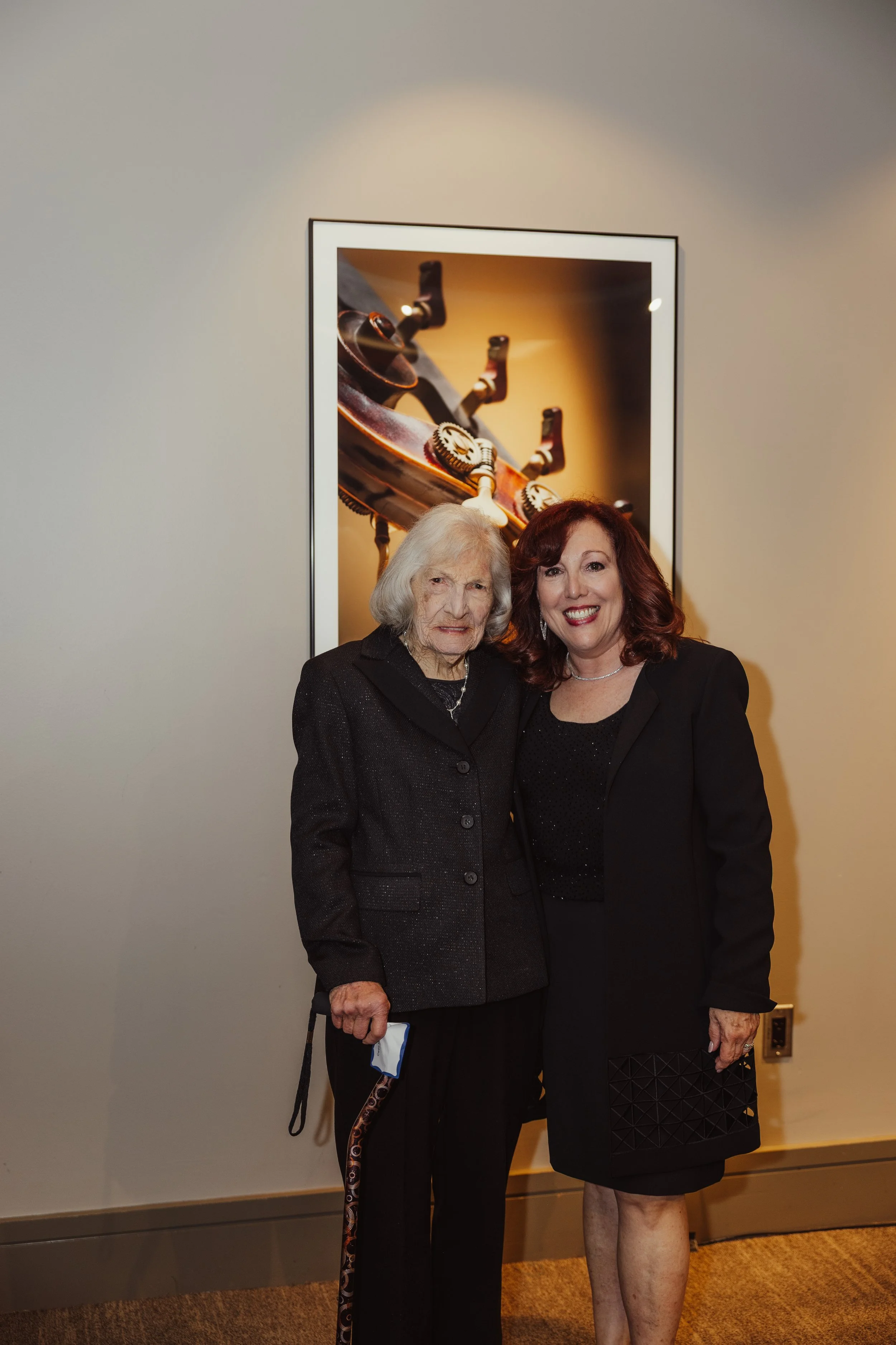 Two women standing together in front of a framed artwork, smiling at the camera, one older with gray hair and the other younger with red hair, dressed in black.