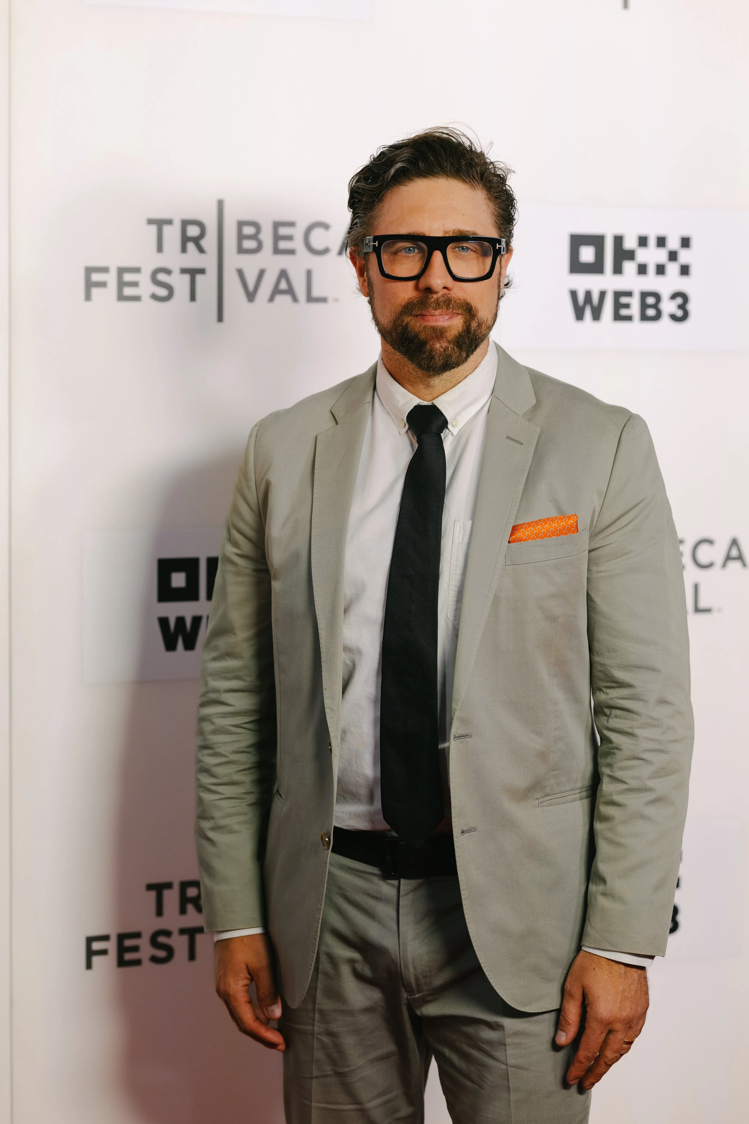 A man in a light gray suit, white shirt, black tie, and black glasses standing in front of a step-and-repeat backdrop with event branding for Tribeca Fest and Web3.